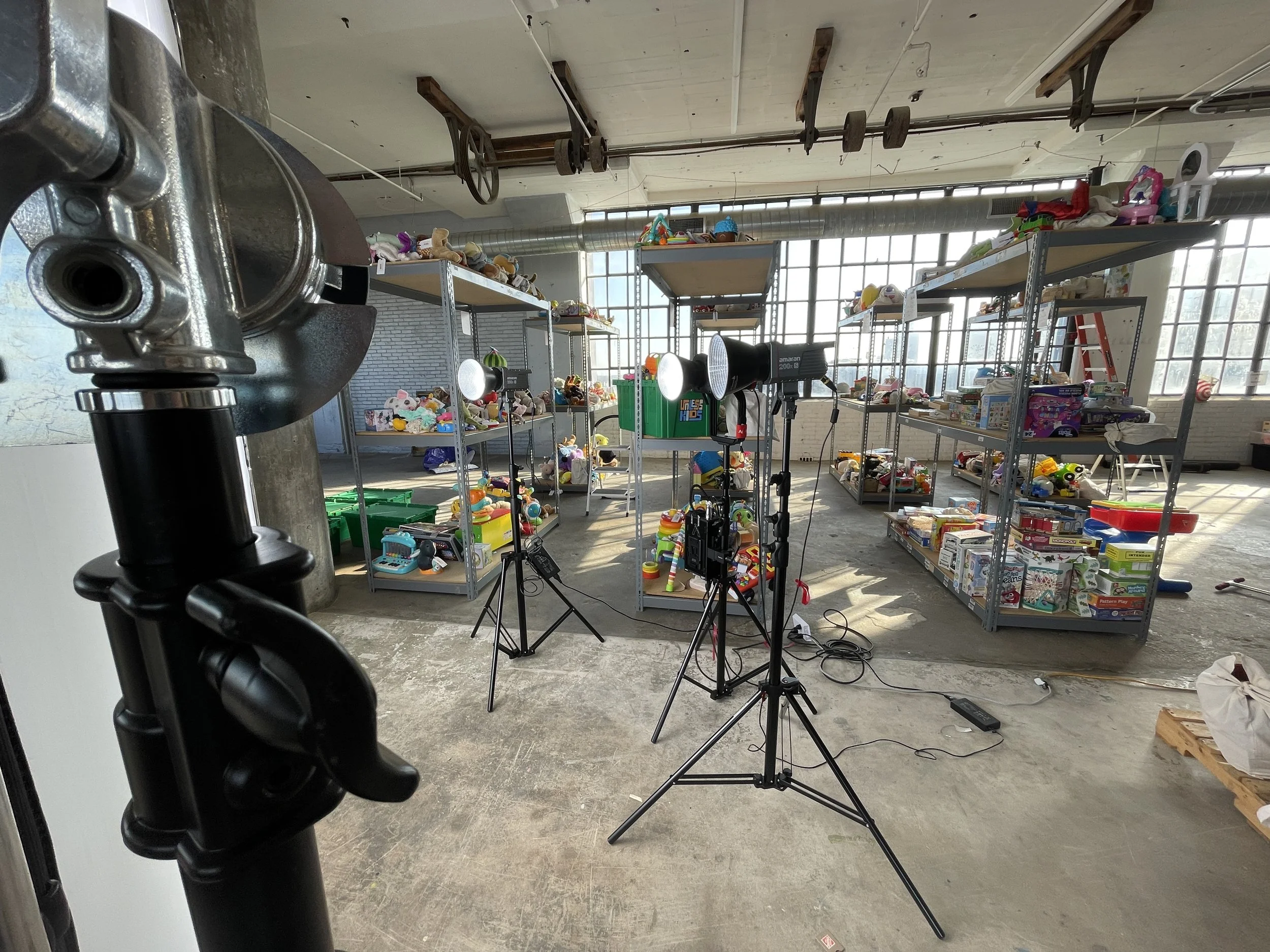 Studio setup with lighting equipment and shelves filled with toys and miscellaneous items, illuminated by large windows.