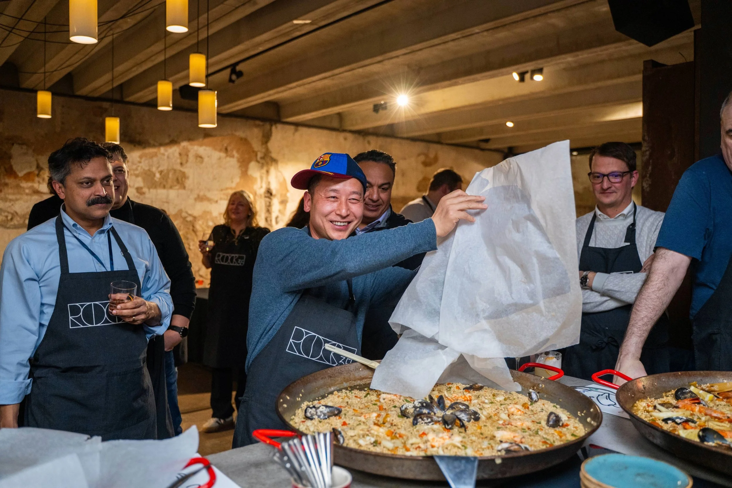 A group of people in a cooking class or event, with a man in the center lifting a paper cover from a large pan of seafood paella, surrounded by others watching and smiling, in a rustic indoor setting with warm lighting.