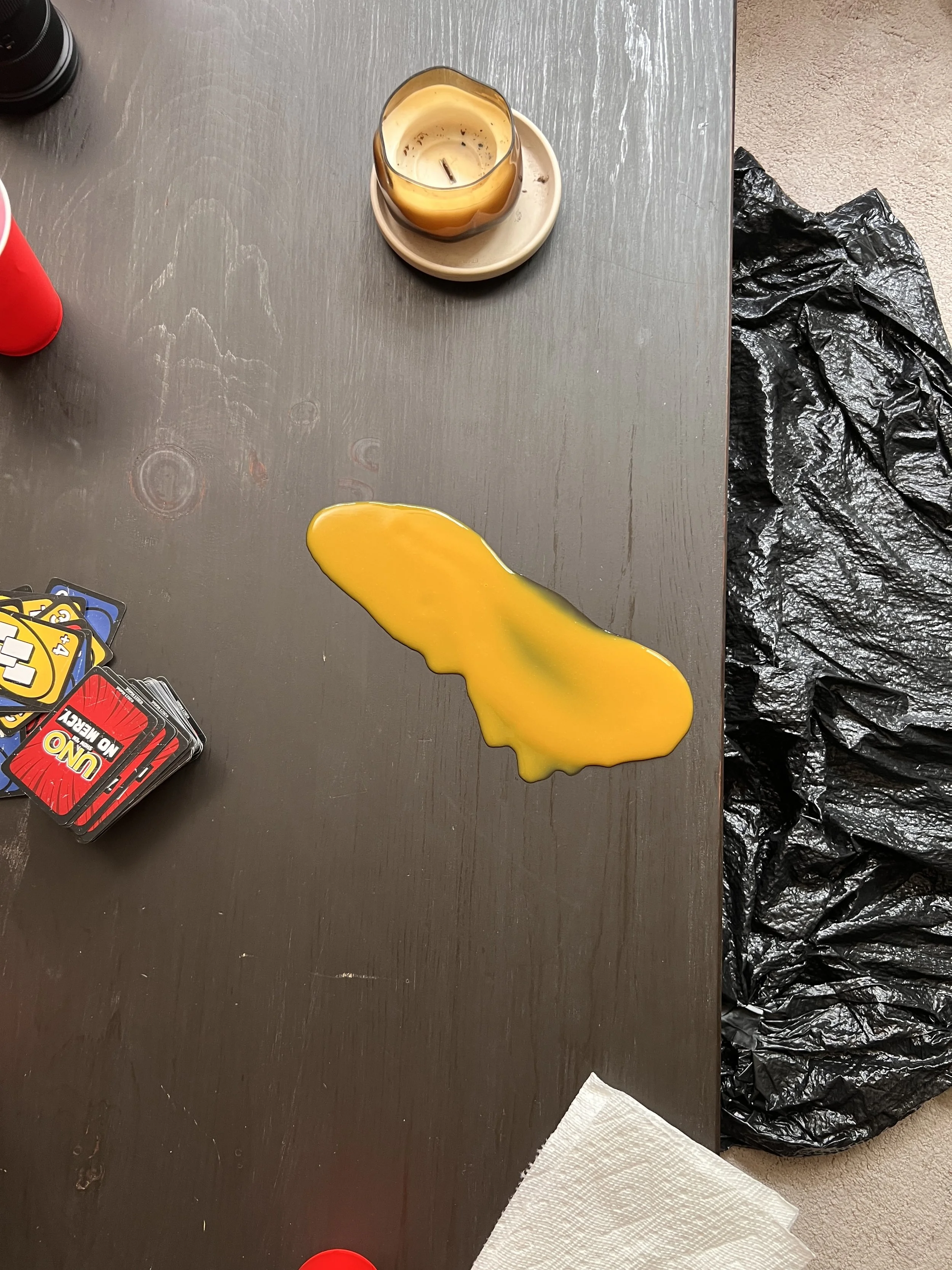 A spilled yellow liquid on a wooden table. There are scattered cards, a candle in a glass holder, and a black plastic bag on the right side.