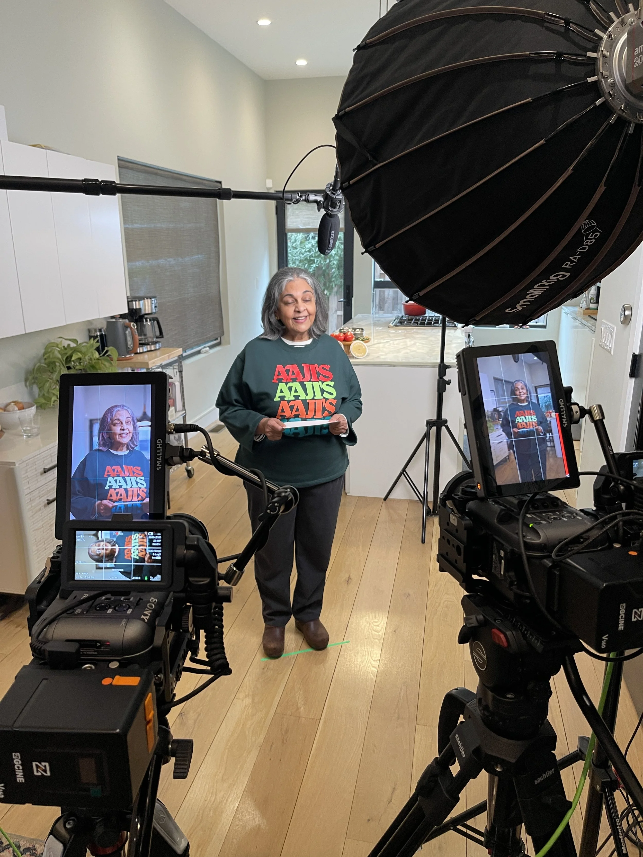 A woman is standing in a kitchen, speaking in front of cameras and professional lighting for a video shoot or interview. She is wearing a dark green sweatshirt with colorful text on it.