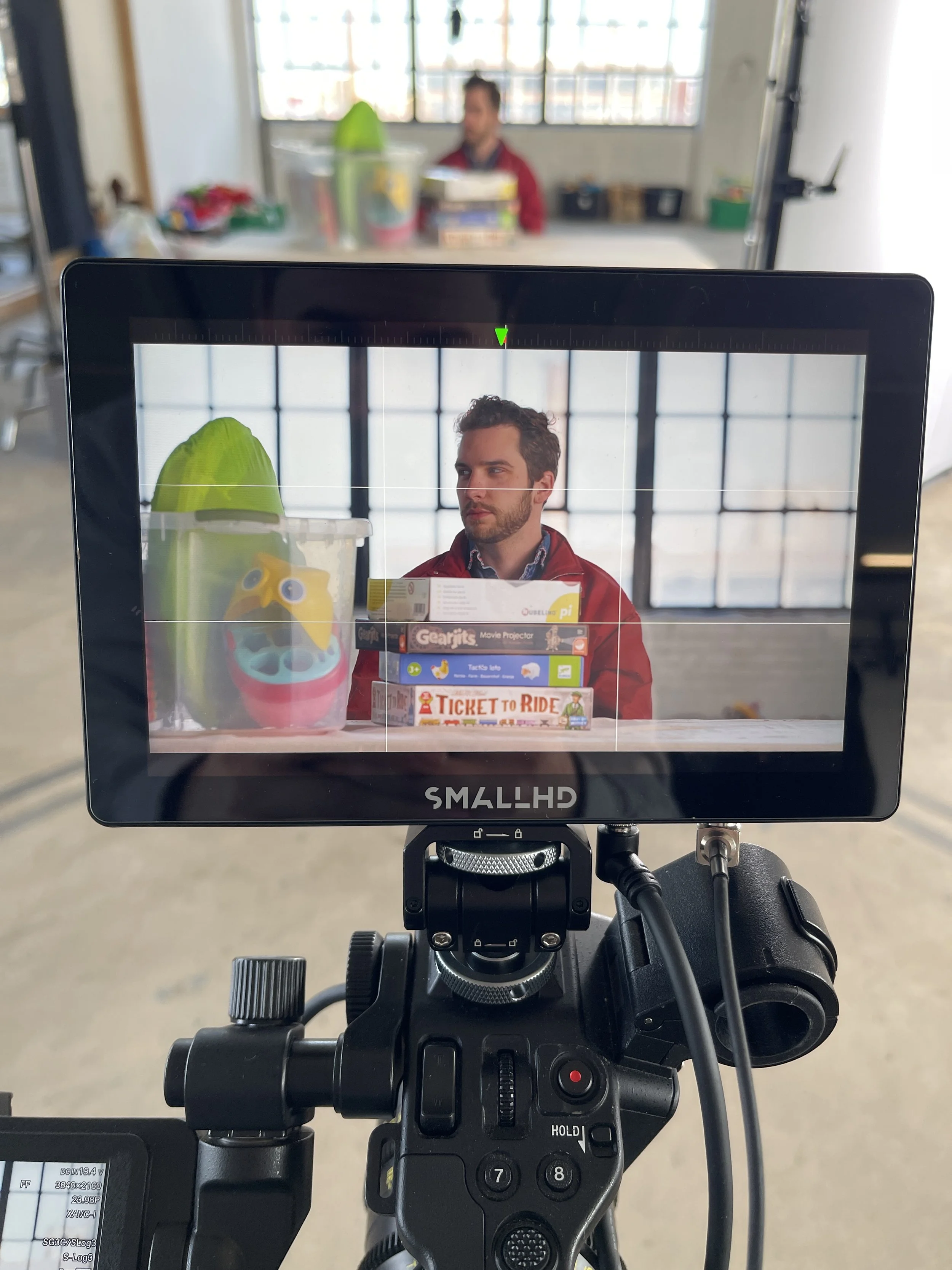 A camera view of a man sitting at a table with stacking board game boxes, with a colorful plastic container shaped like a character and large purple sunglasses on it, and a green object in the background.