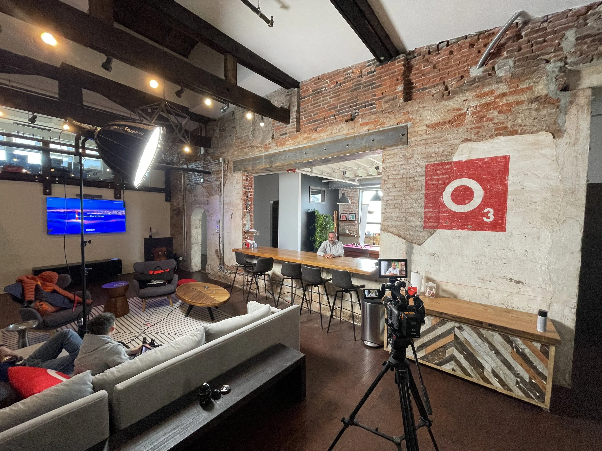 Interior of a modern loft-style room with exposed brick and concrete walls, featuring a bar area with black chairs, a large television, a sofa, a camera on a tripod, and people sitting and preparing for filming.