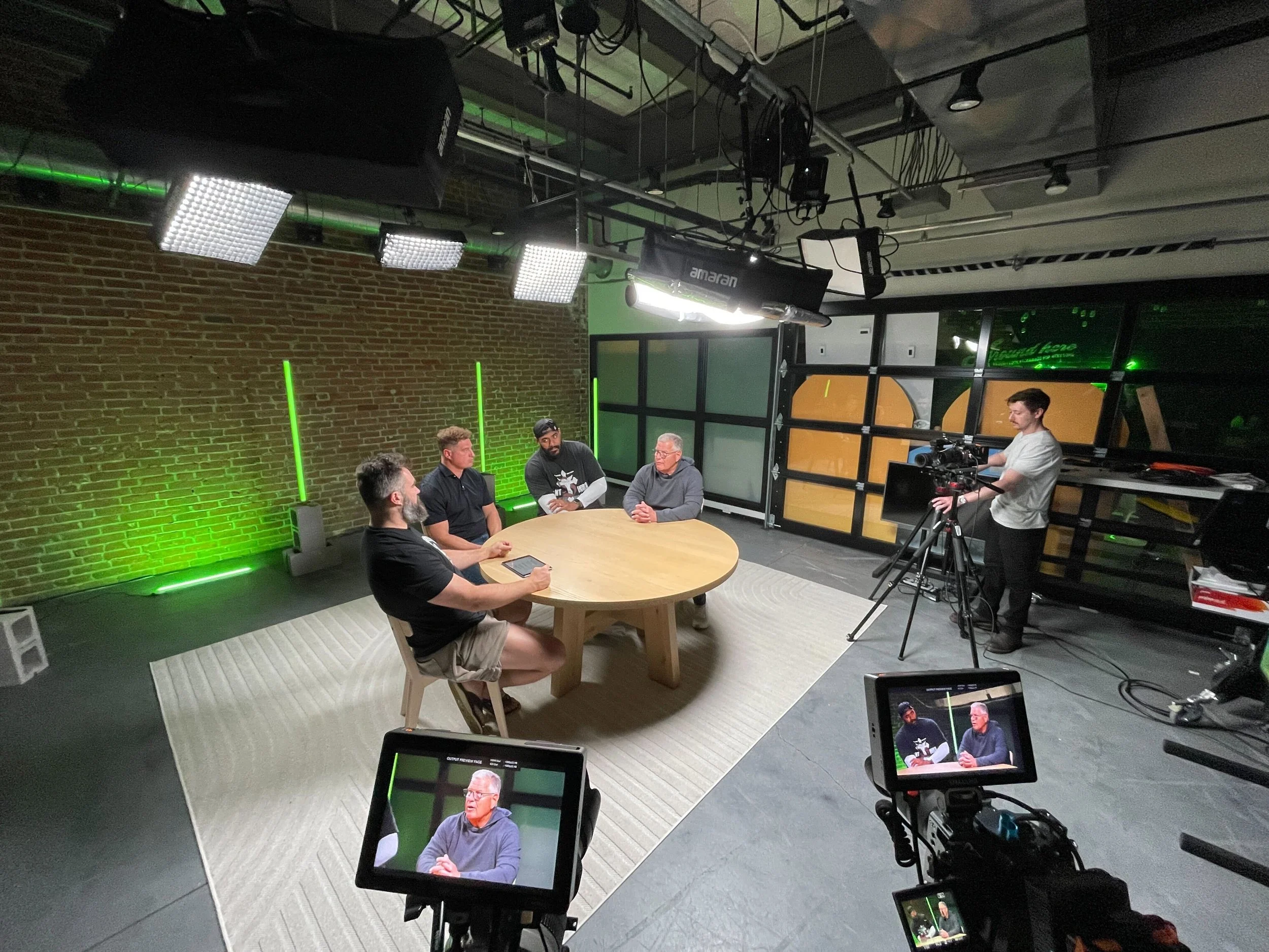 A behind-the-scenes view of a recording or interview session in a studio with several people seated at a round table, a cameraman filming, studio lights hanging from the ceiling, and monitors showing a man speaking during the interview.