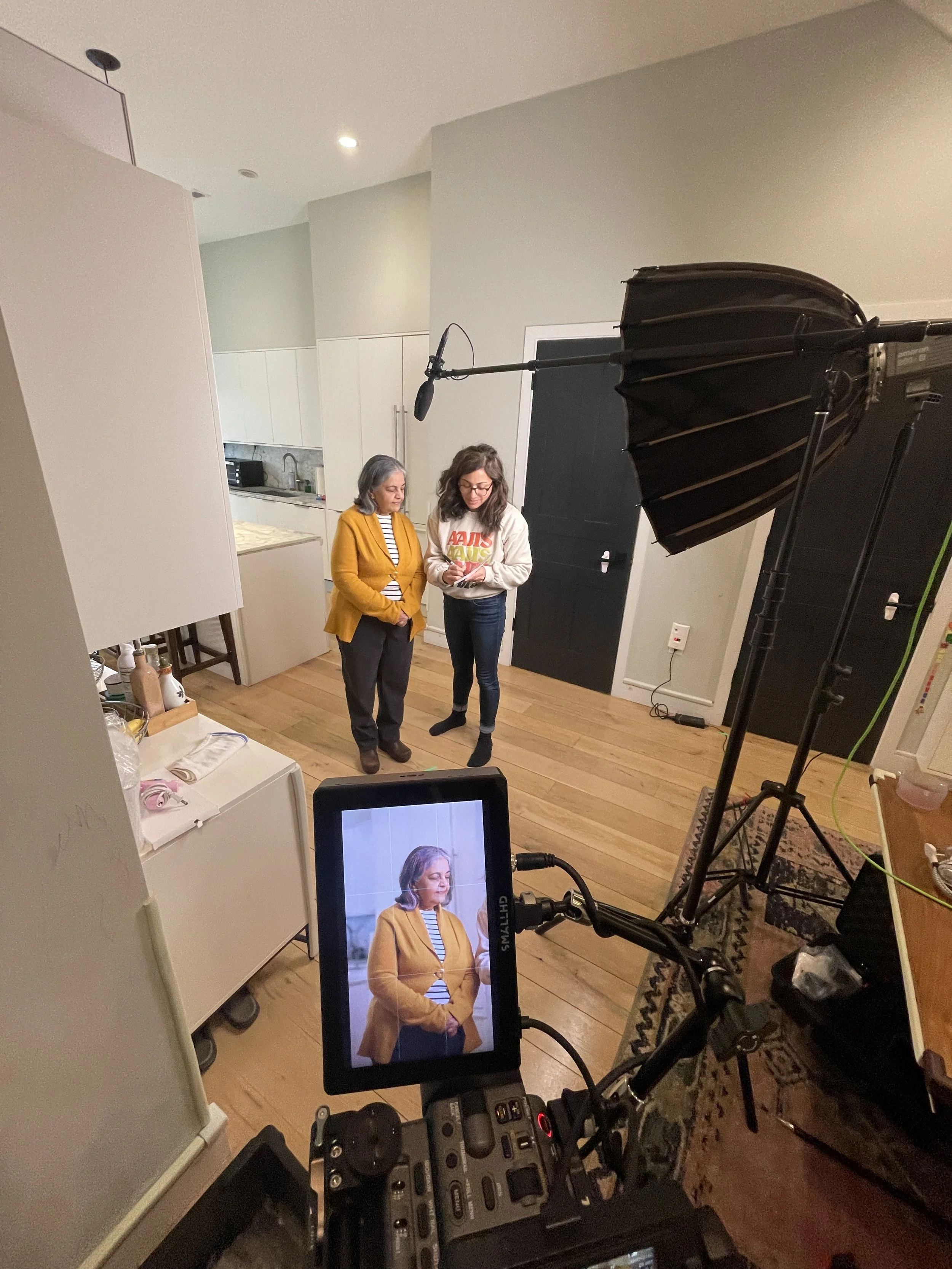 A behind-the-scenes shot of a video or photo shoot inside a home, with a camera on a tripod capturing two women standing together, one with gray hair and a mustard yellow sweater, and the other with brown hair and glasses, wearing a white sweatshirt. Equipment includes a large studio light and a monitor showing the image being recorded.