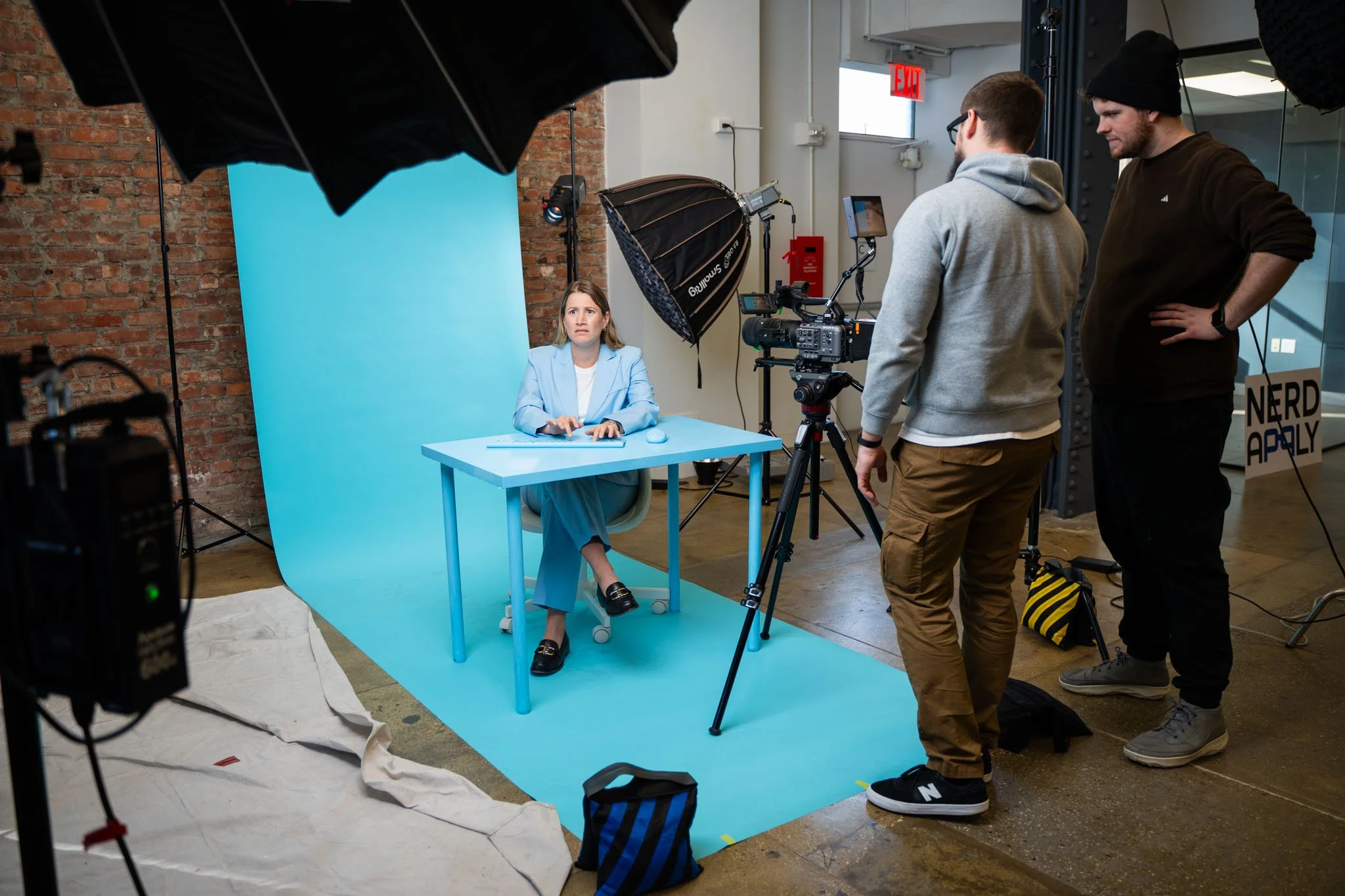 A woman sits at a blue desk in a photo studio with a blue backdrop, filming a video. Two men operate camera equipment, one standing behind the camera and the other to the side. Studio lighting and equipment are visible, with a brick wall and an emergency exit door in the background.