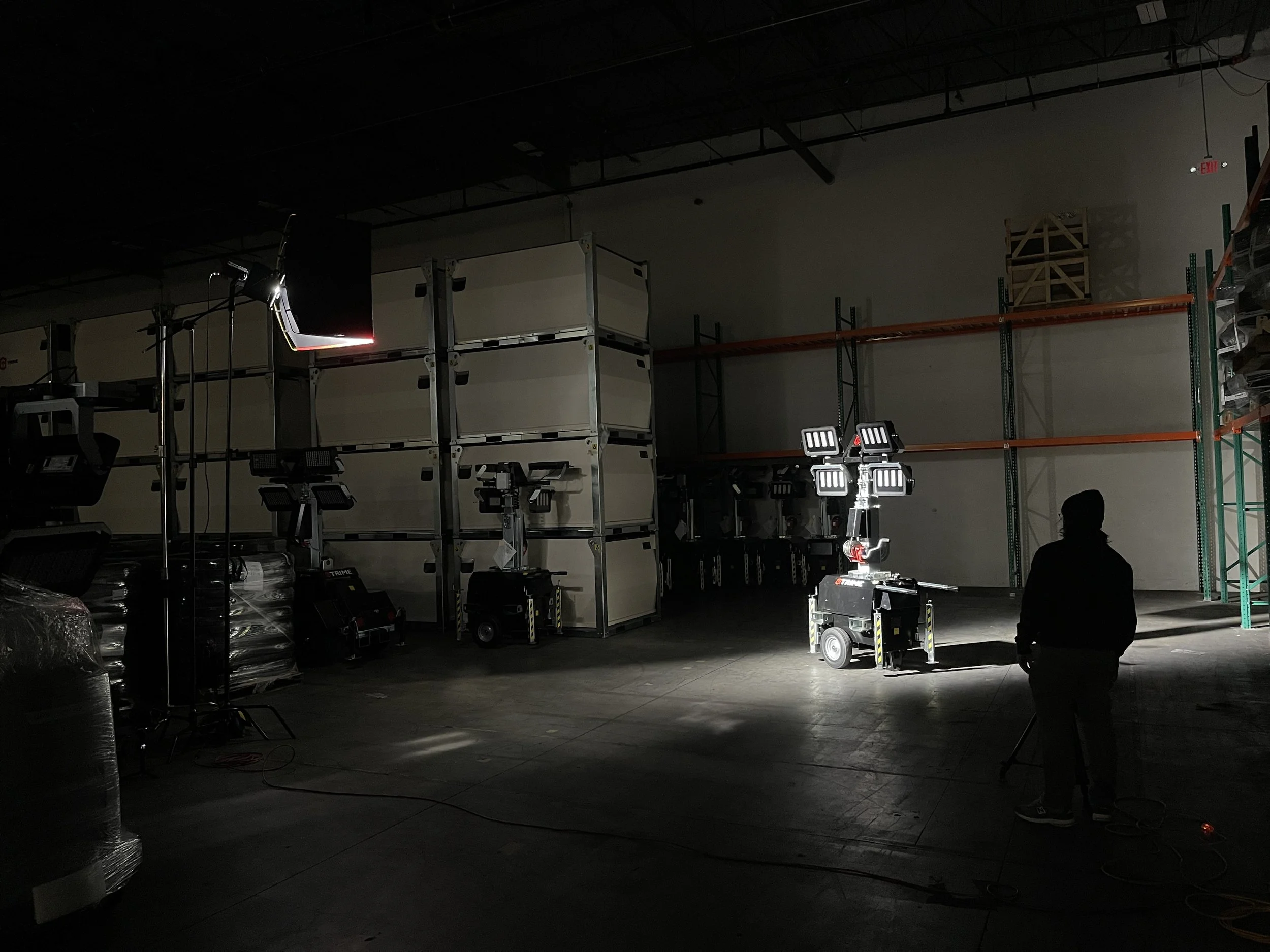 A person stands in a dark warehouse with stage lighting equipment and scaffolding. Several LED lights and portable power sources are visible on the floor.