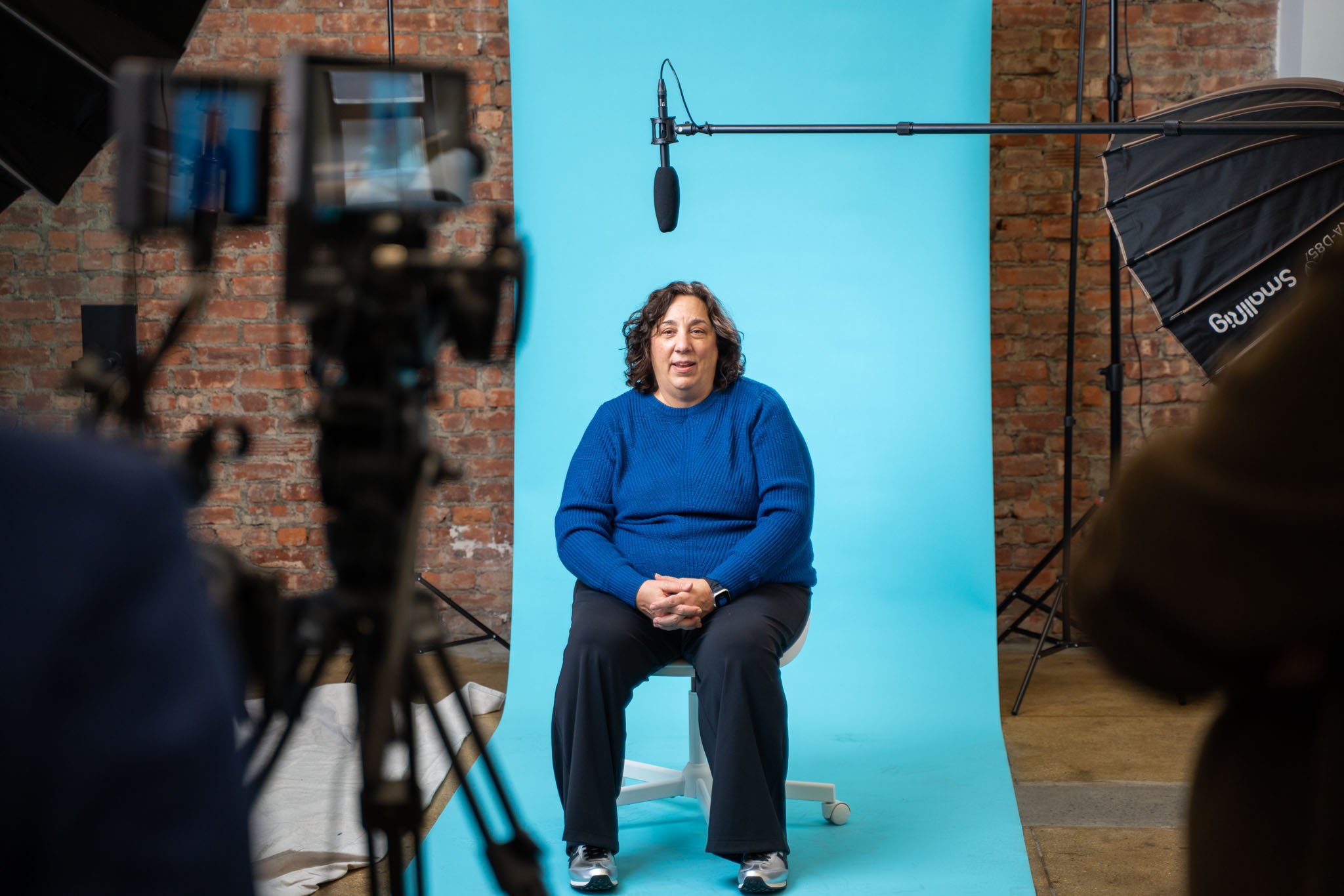 Woman sitting on a chair in front of a blue backdrop during a video or photo shoot, with professional lighting and camera equipment around.