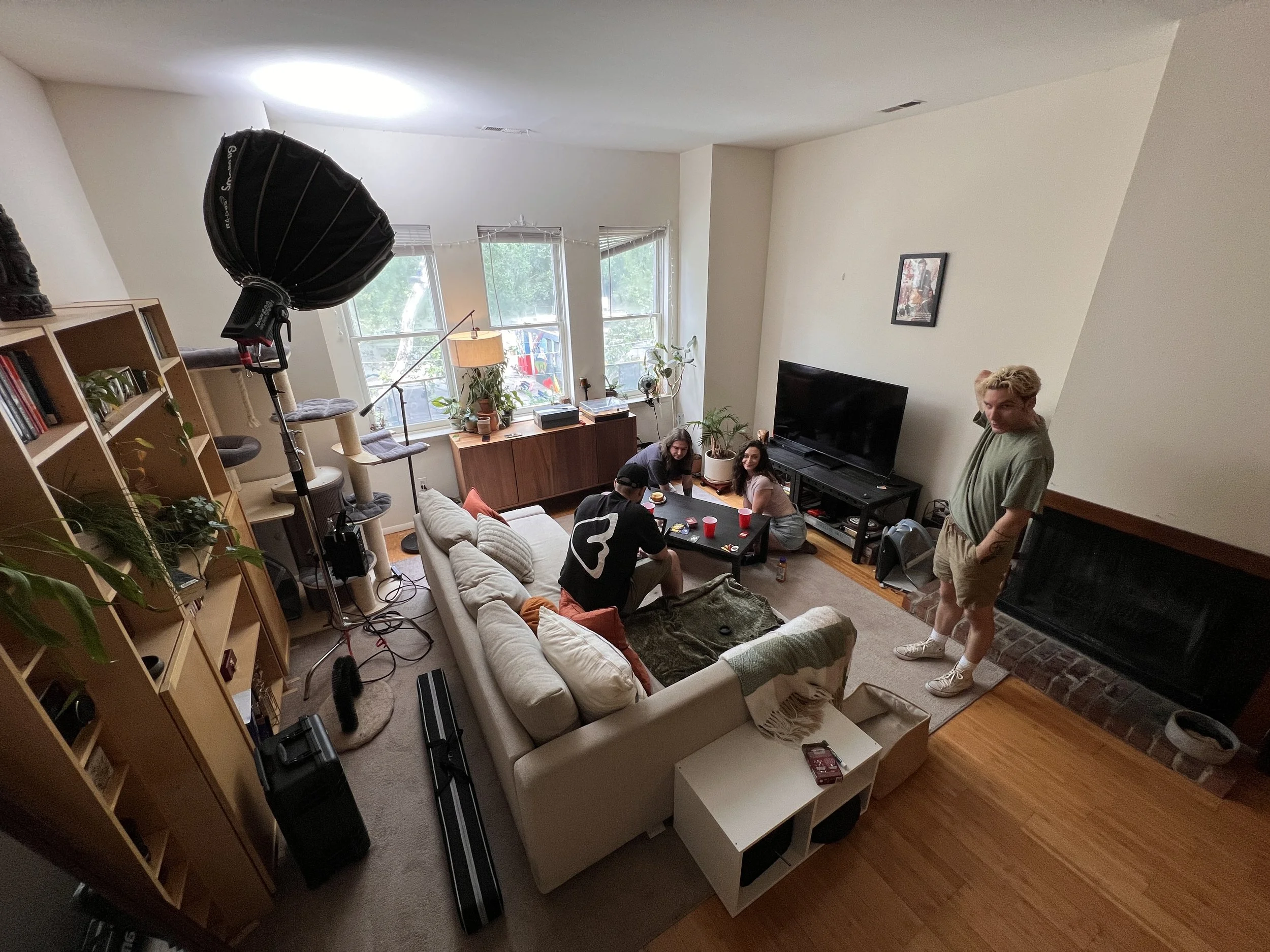 Living room with several people, large window, TV, bookshelf, plants, and a person standing near the fireplace.