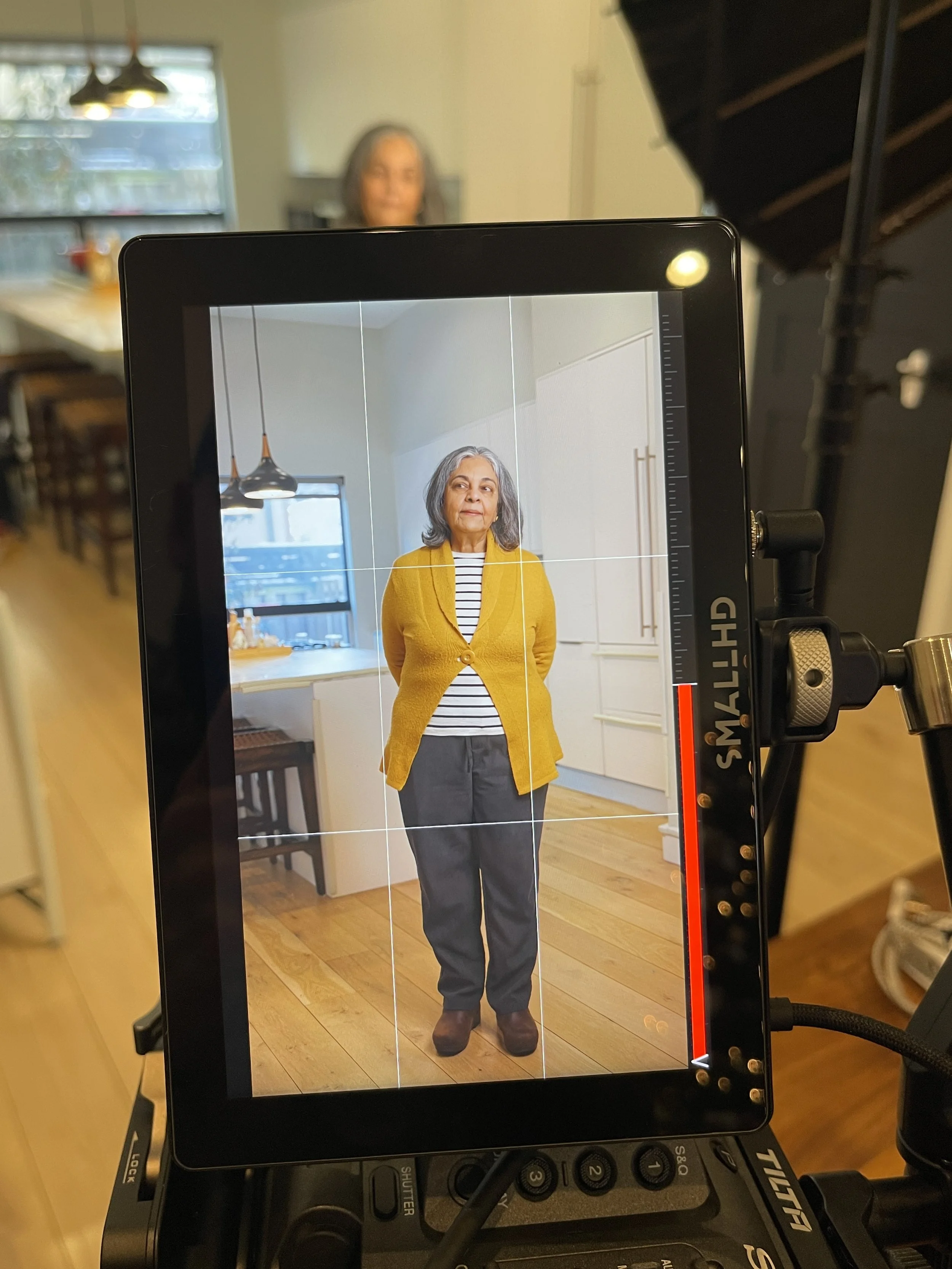 An elderly woman wearing a yellow cardigan, striped shirt, gray pants, and brown shoes standing in a bright kitchen, during a photo shoot.