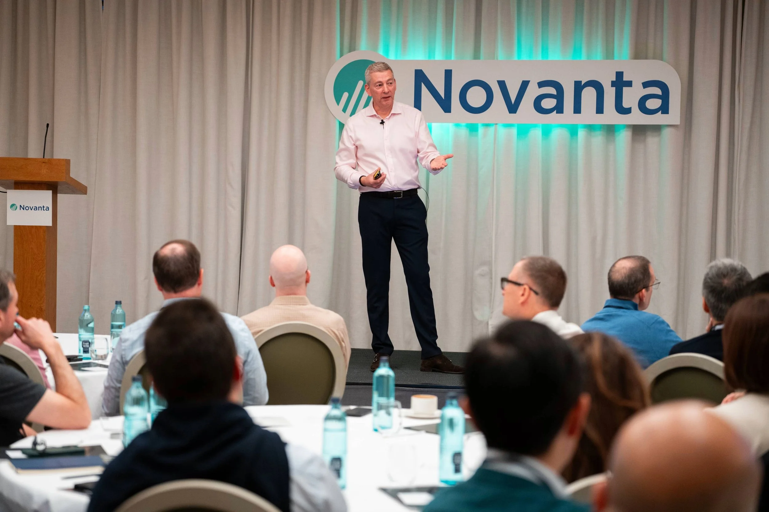 A man in a white shirt and black pants giving a presentation on stage at a Novanta conference, with a seated audience listening.