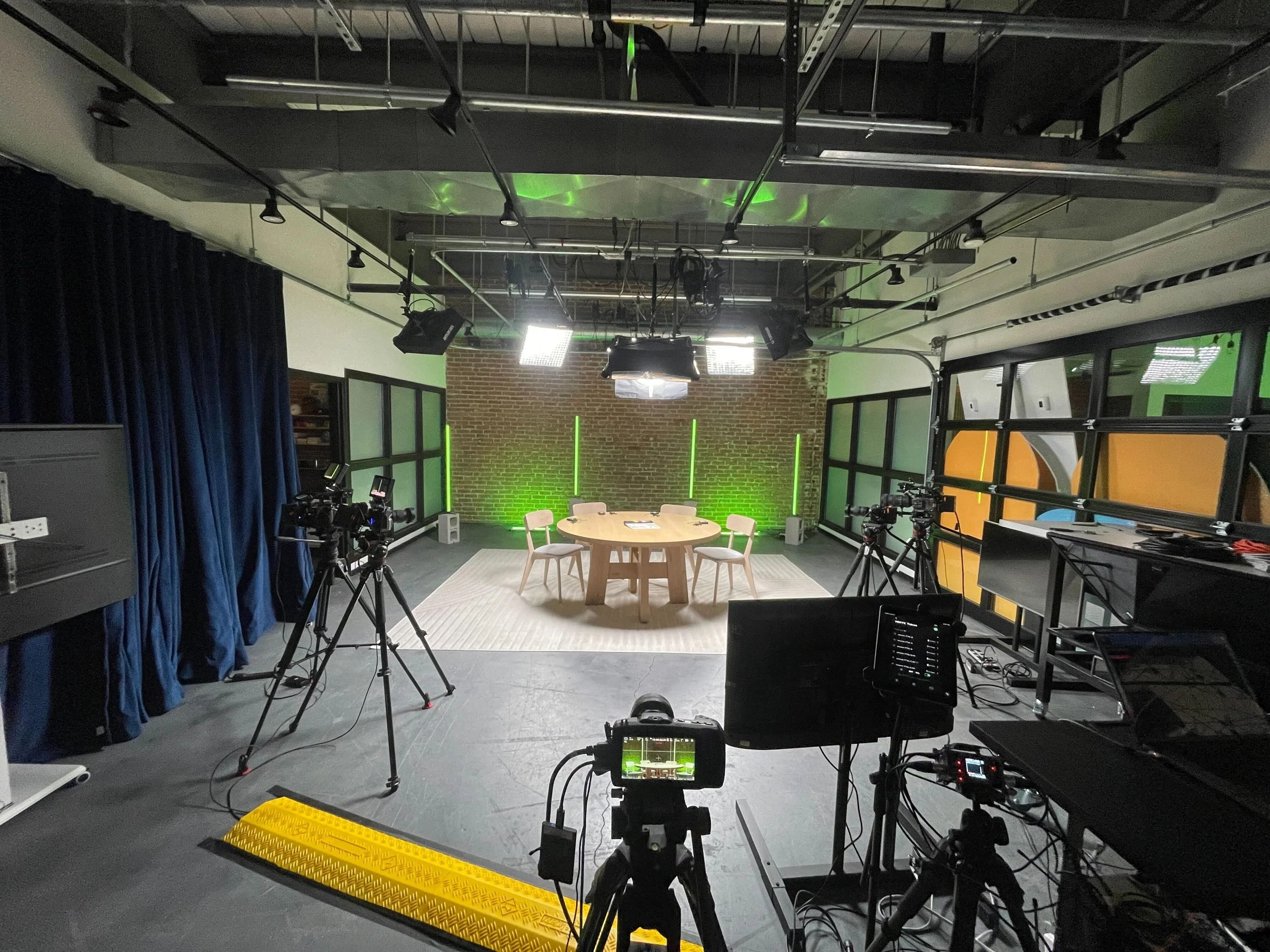 The image shows a television or film studio with filming equipment, including cameras on tripods, lighting, and monitors. There is a round table with chairs in the center, positioned on a light-colored rug, against a background of exposed brick wall with green lighting accents and glass window panels.