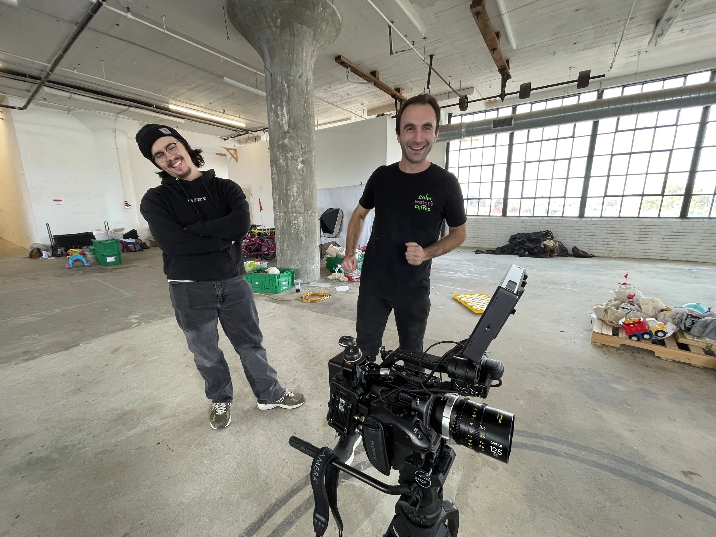Two men smiling and posing in an industrial-style space with large windows and construction materials. One man wears a black hoodie and beanie, the other wears a black t-shirt. A professional camera is set up in the foreground.