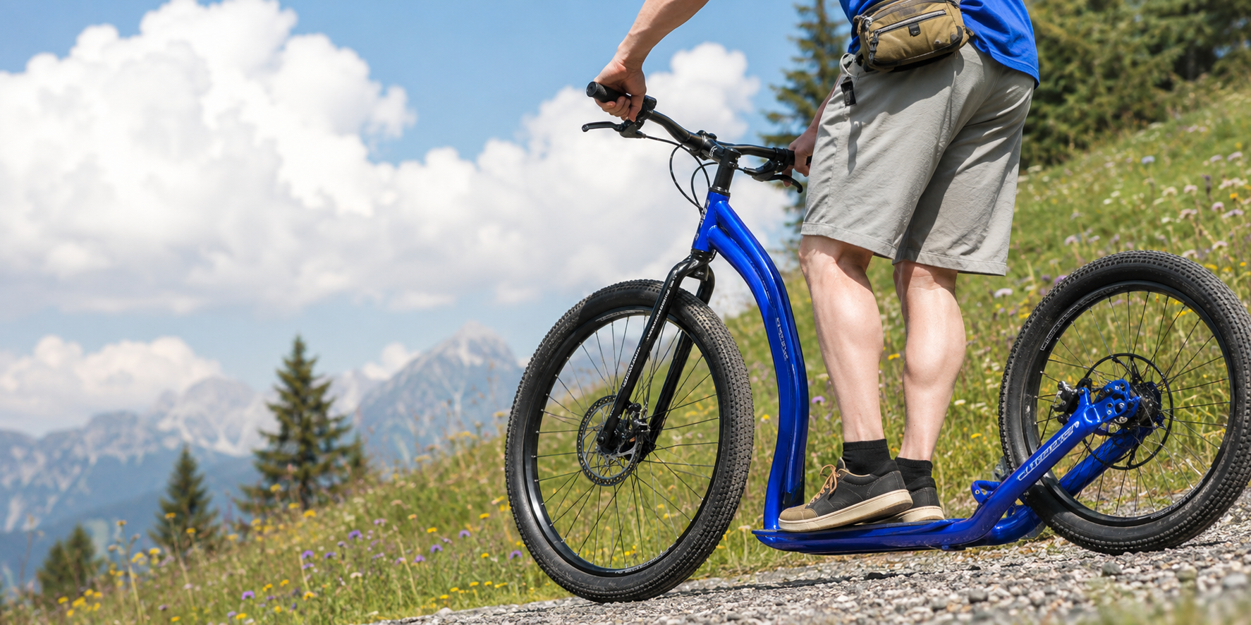Person standing on a blue electric scooter on a gravel trail in a scenic mountainous area with trees and flowering plants, wearing shorts, a blue shirt, and a small bag.