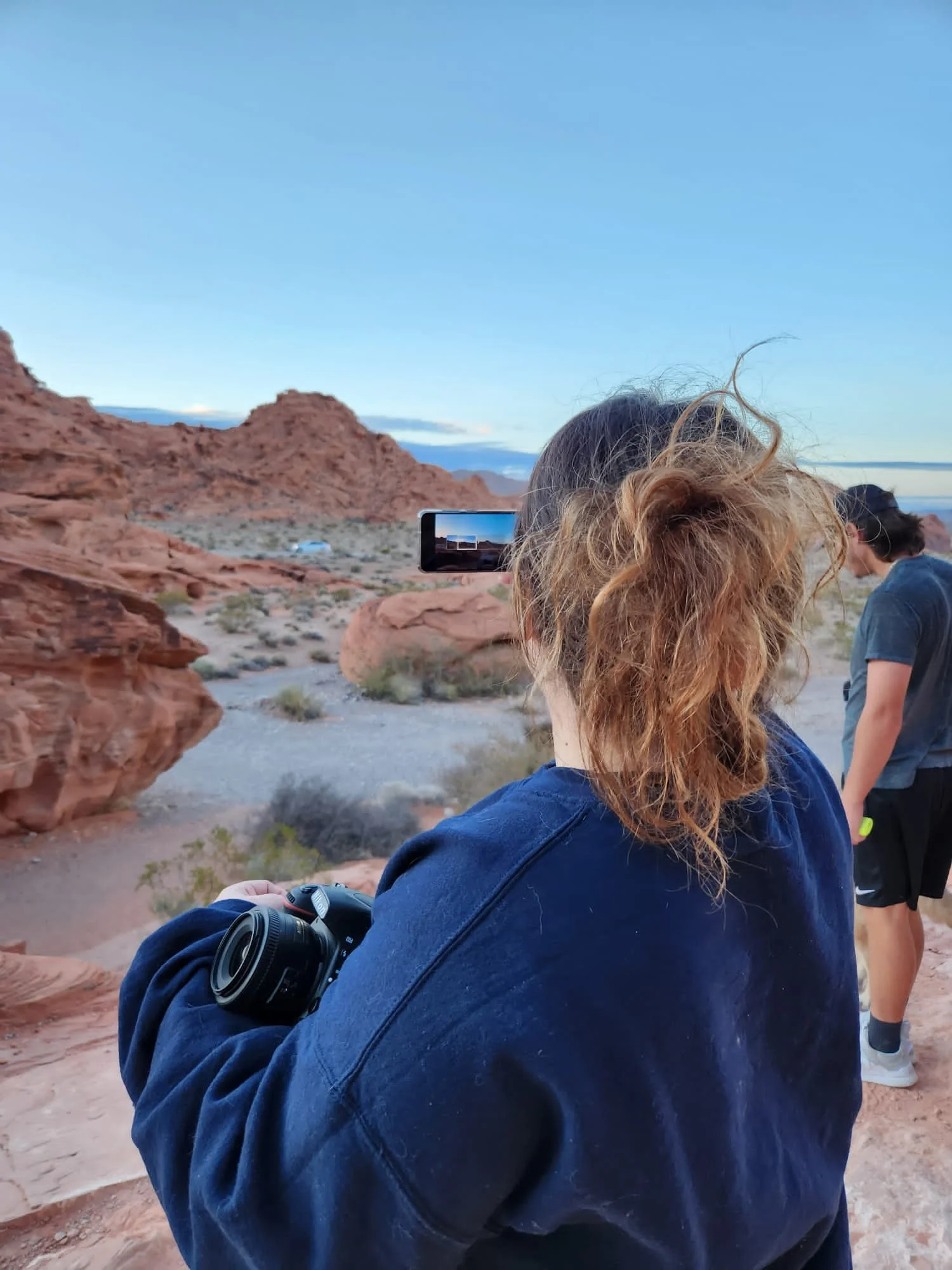 A woman with curly red hair taking a photo of a desert landscape with a smartphone, in a rocky, red terrain with mountains in the background, with another woman nearby.