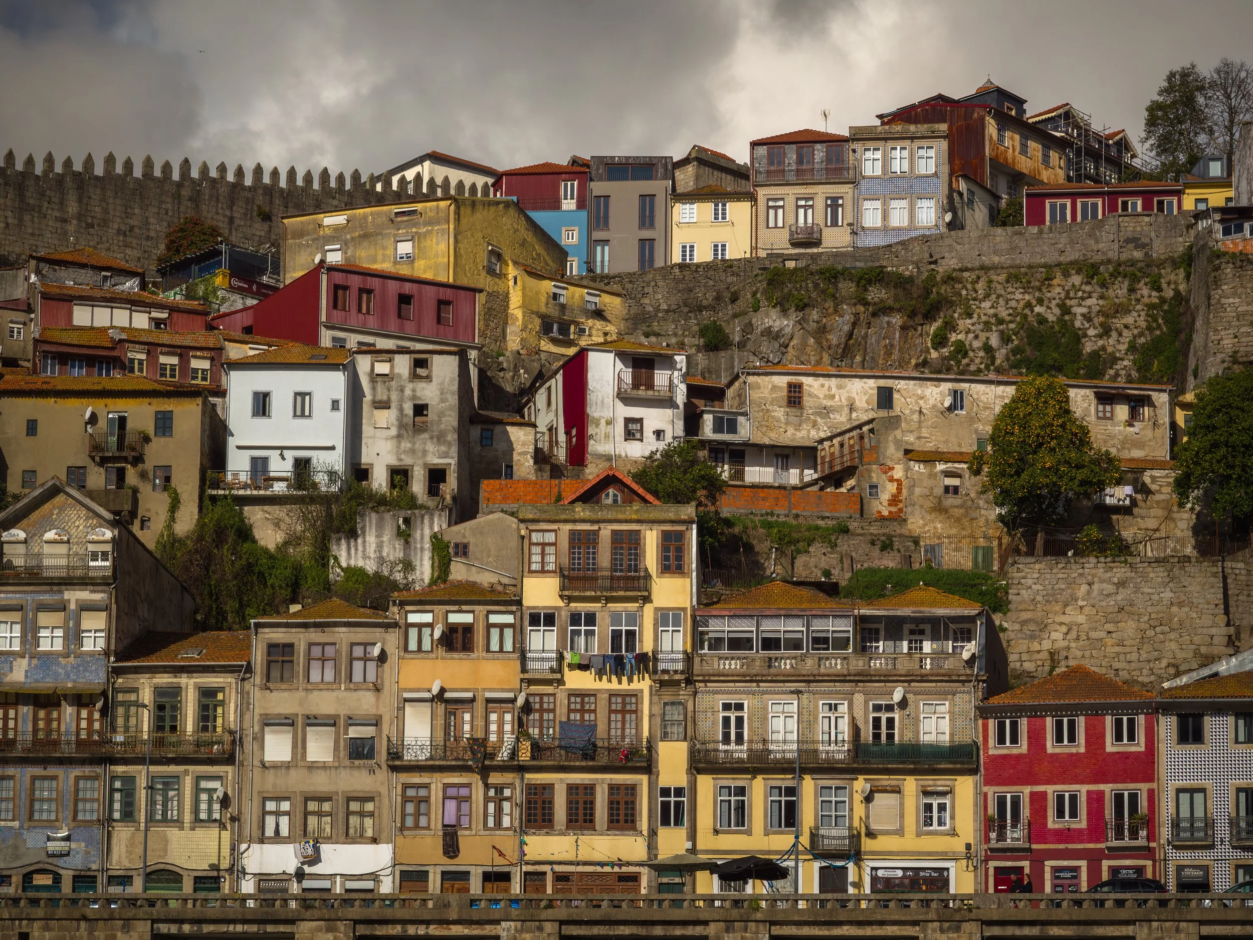 Une rangée de bâtiments colorés et variés en contrebas d'une colline escarpée, avec un mur de ville en pierre au sommet, dans un environnement urbain.