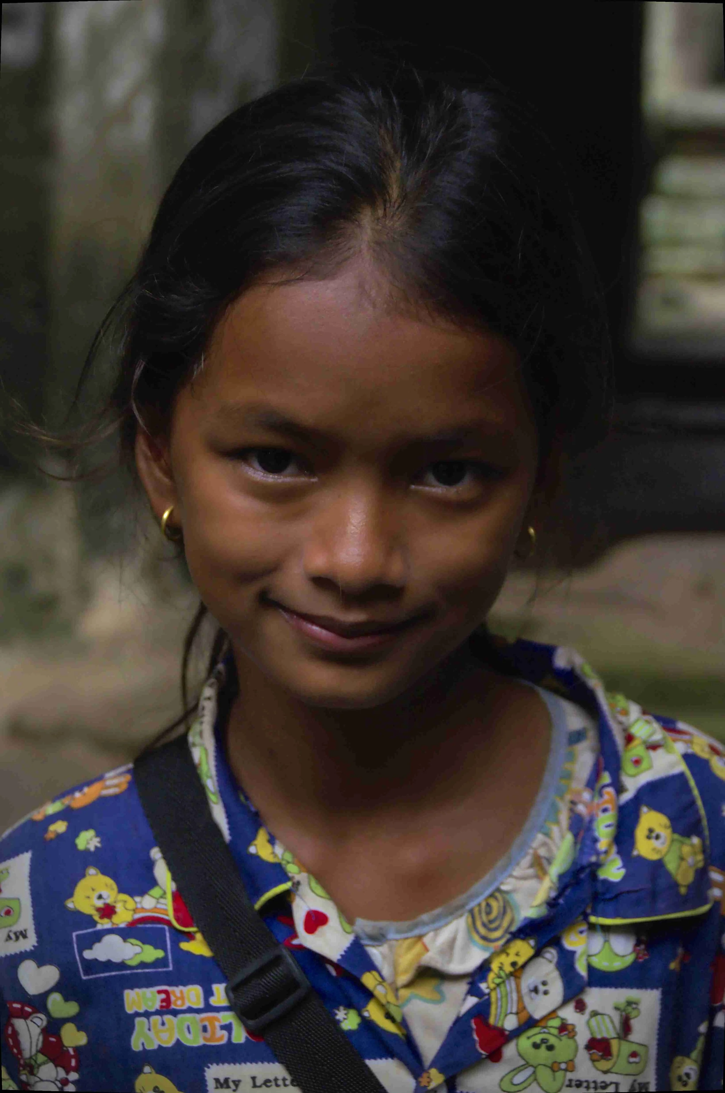Une jeune fille souriante avec de longs cheveux noirs, portant une chemise colorée avec des motifs de personnages de dessins animés, et une petite boucle d'oreille en or.