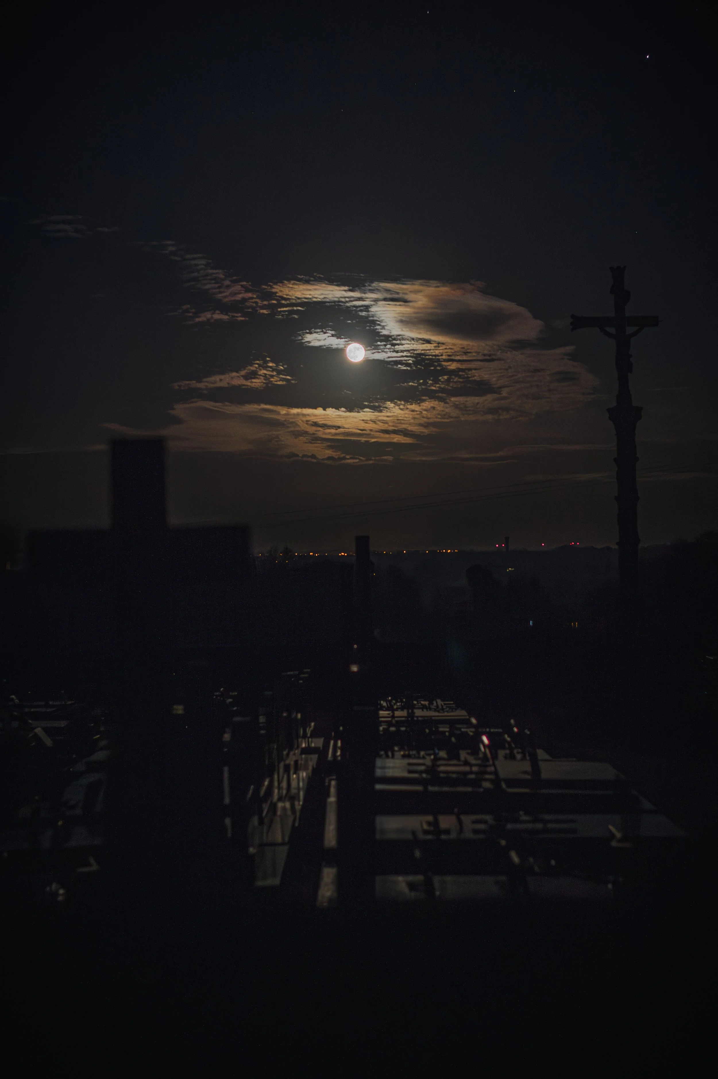 Paysage nocturne avec la lune partiellement couverte par des nuages, silhouette d'une croix en bois au premier plan.