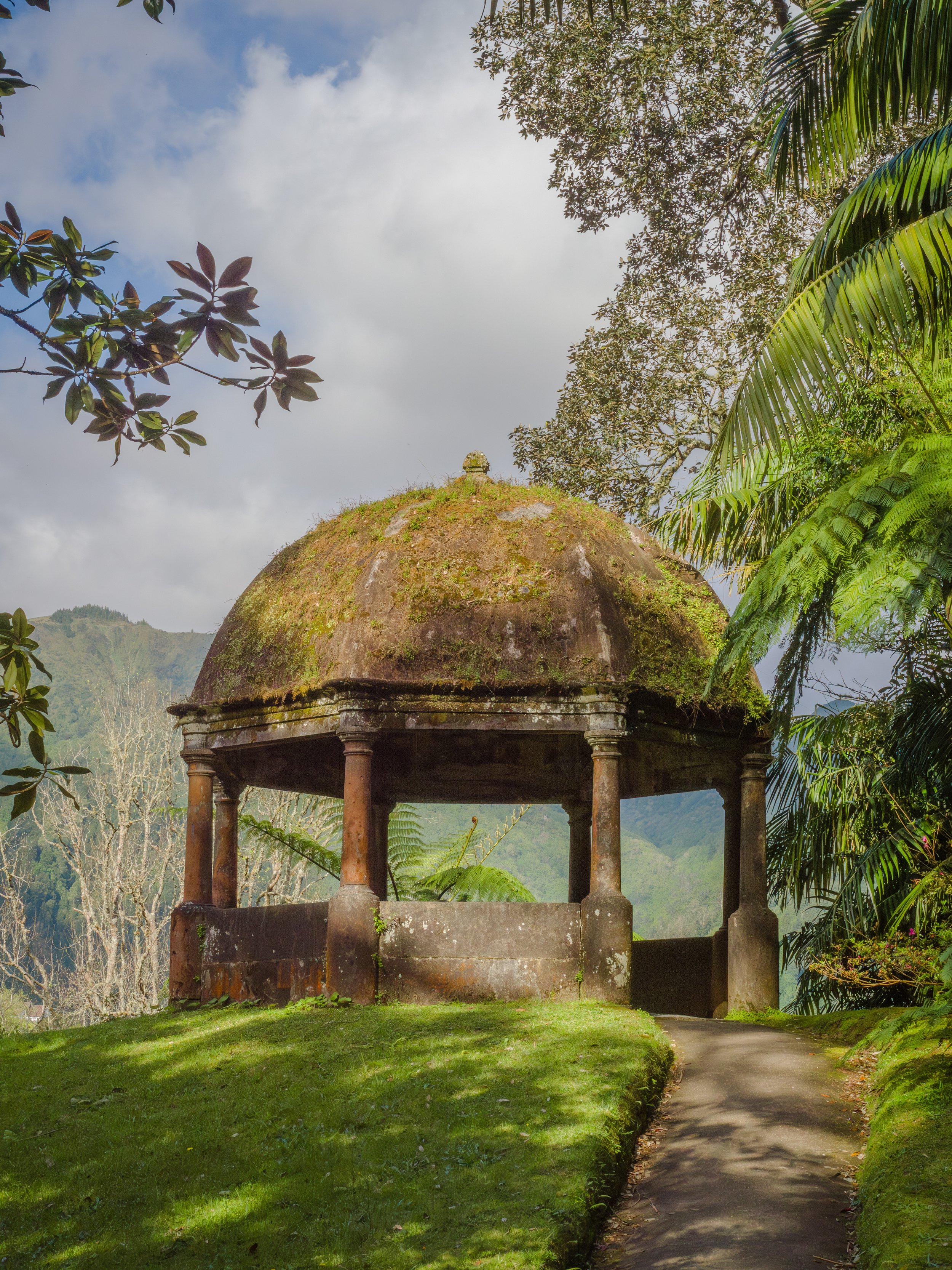 Une petite structure en pierre avec un toit en dôme recouvert de mousse, entourée de végétation dense, sous un ciel partiellement nuageux.