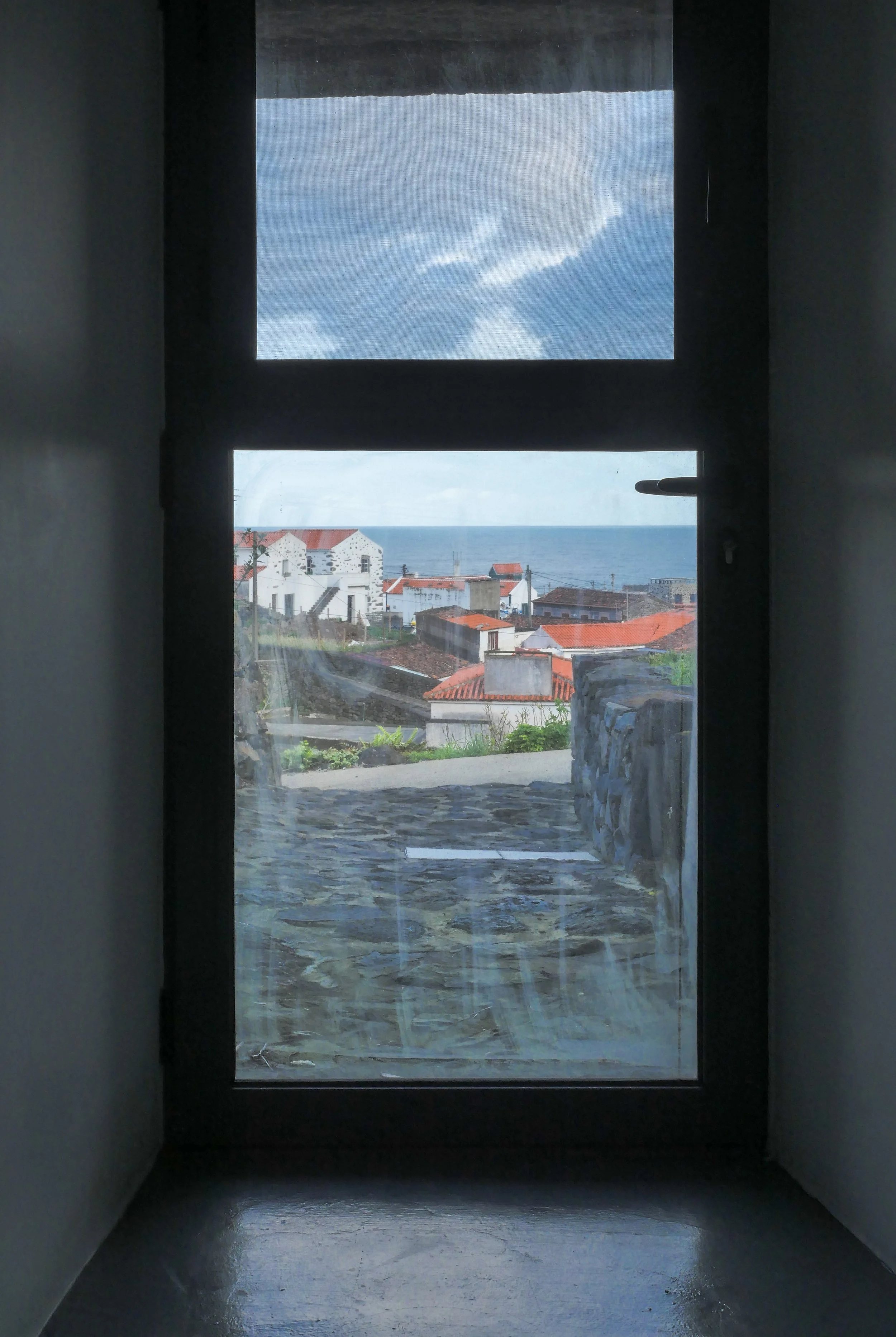 Vue d'une fenêtre avec vue sur des bâtiments avec toits en tuiles rouges, la mer et un ciel partiellement nuageux.