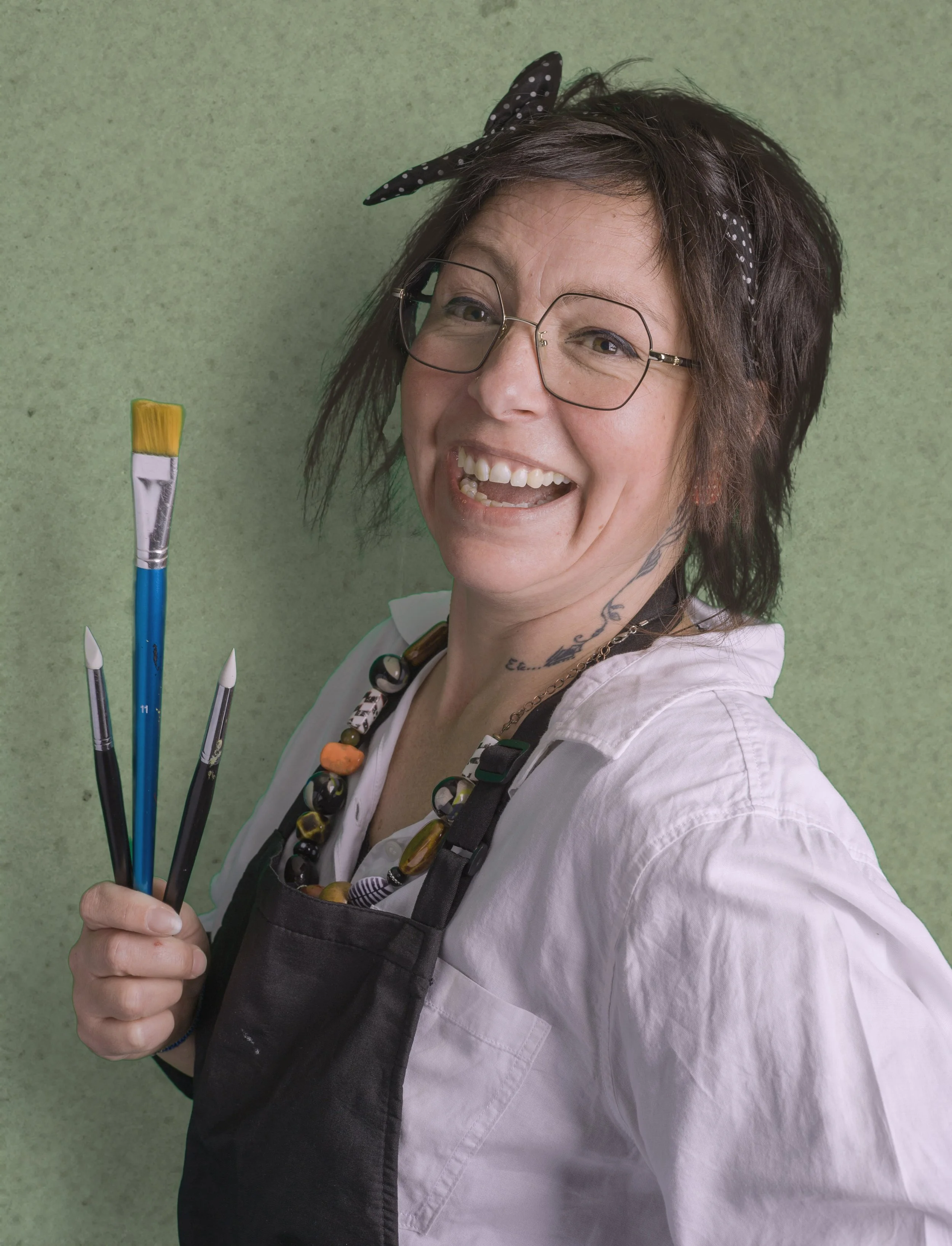 Une femme souriante portant des lunettes, un tablier noir, une chaîne de collier colorée, et tenant trois pinceaux, avec un fond vert.