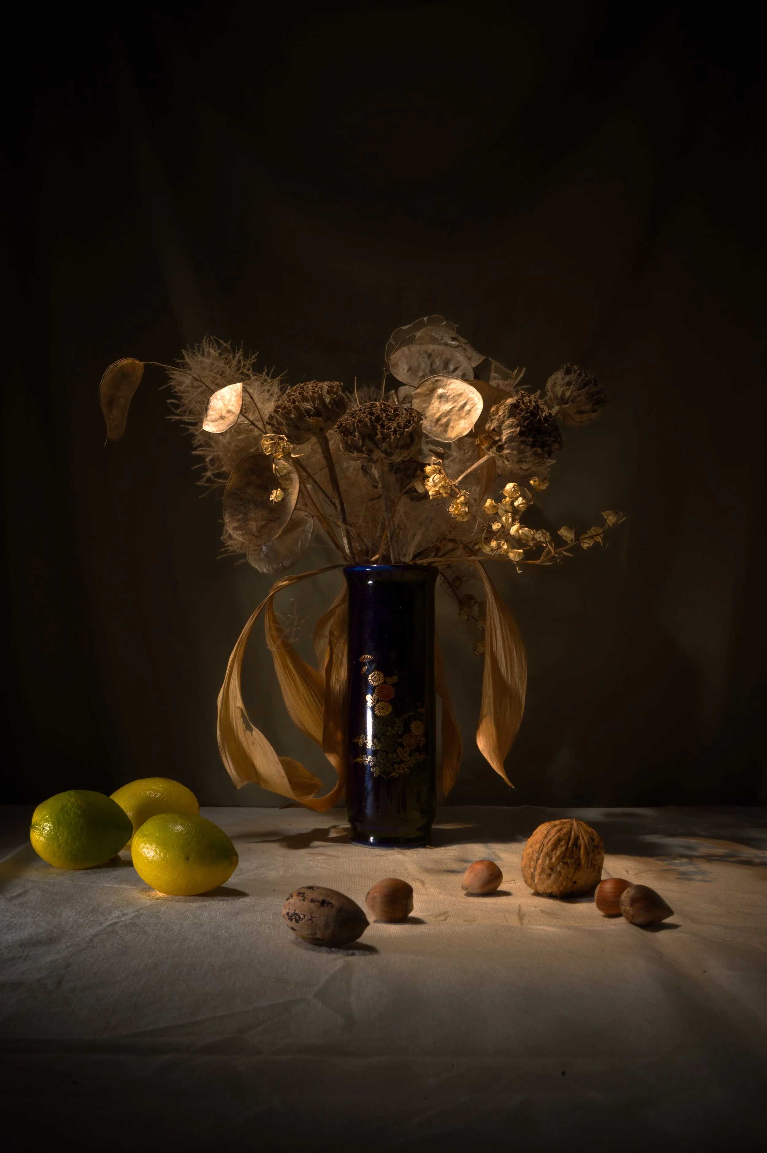Vase noir avec fleurs séchées et fruits à proximité, dans une lumière tamisée.
