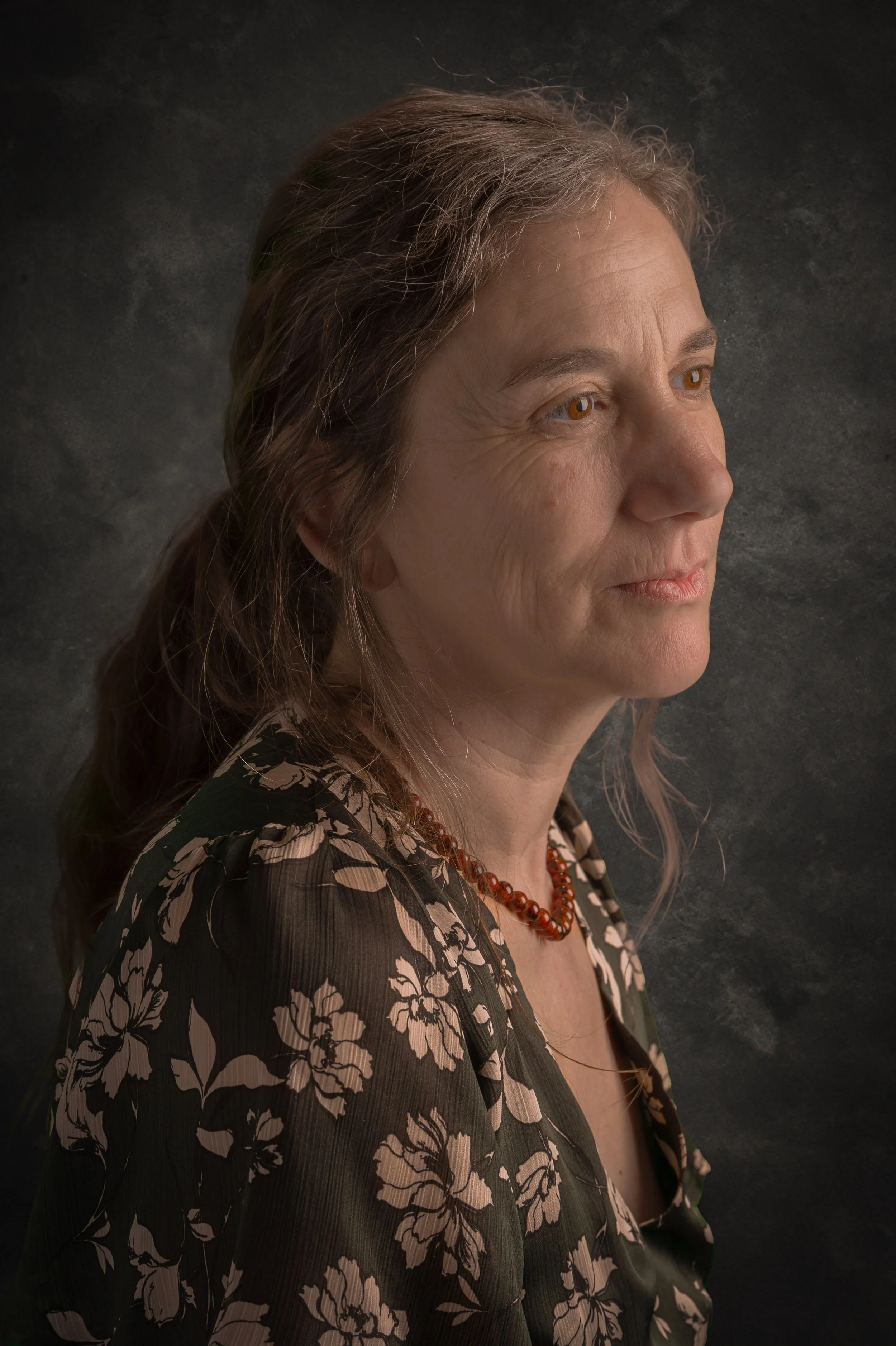 Portrait d'une femme d'âge moyen avec des cheveux gris, portant un chemisier à fleurs noir et beige et un collier en ambre, regardant vers la droite, sur un fond sombre.