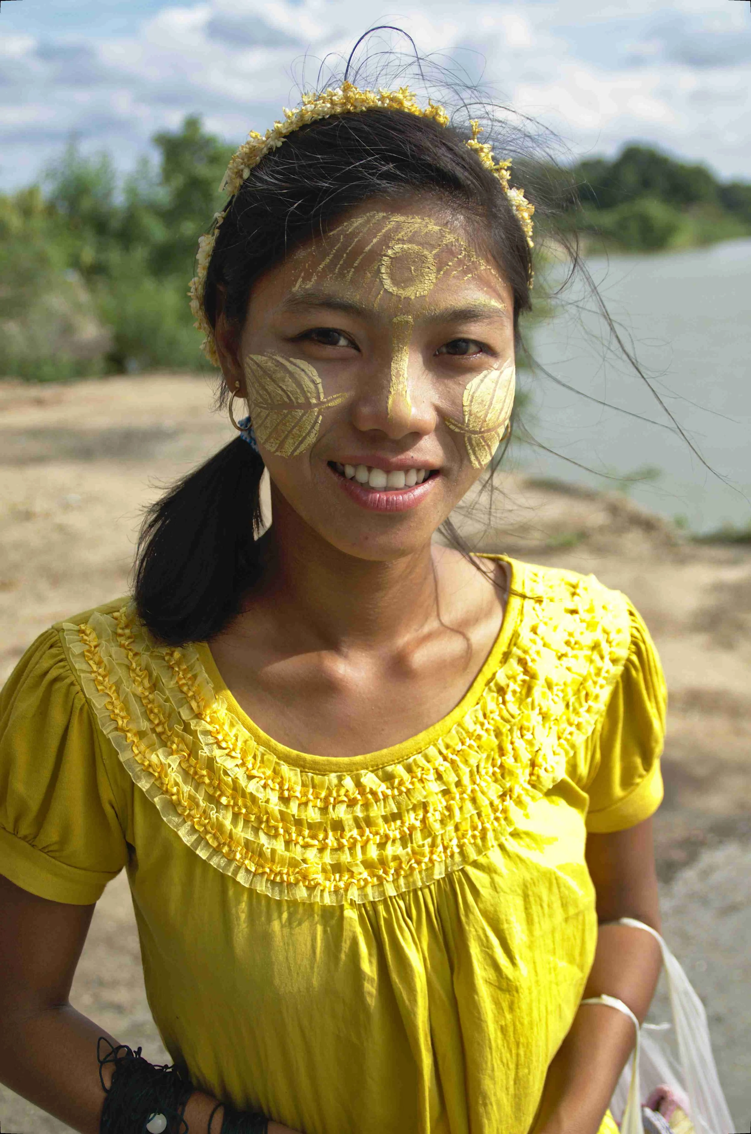 Femme souriante portant une maquillage doré sur le visage, avec un collier et une robe jaune, debout près d'une rivière en plein air.