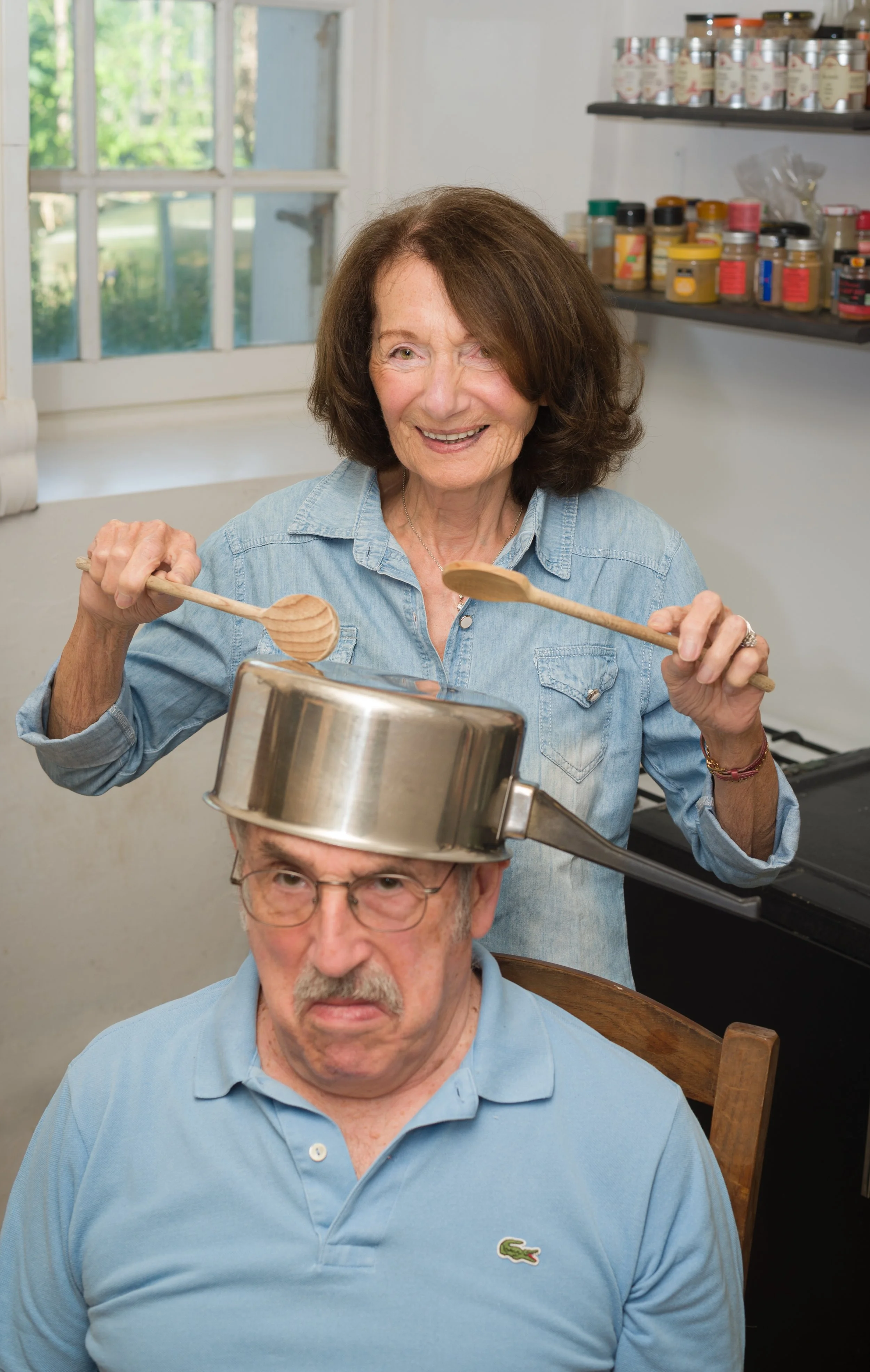 Une femme souriante, tenant deux cuillères en bois, joue avec un homme qui a une casserole sur sa tête et un regard mécontent dans une cuisine. Il porte des lunettes et un polo bleu Lacoste.