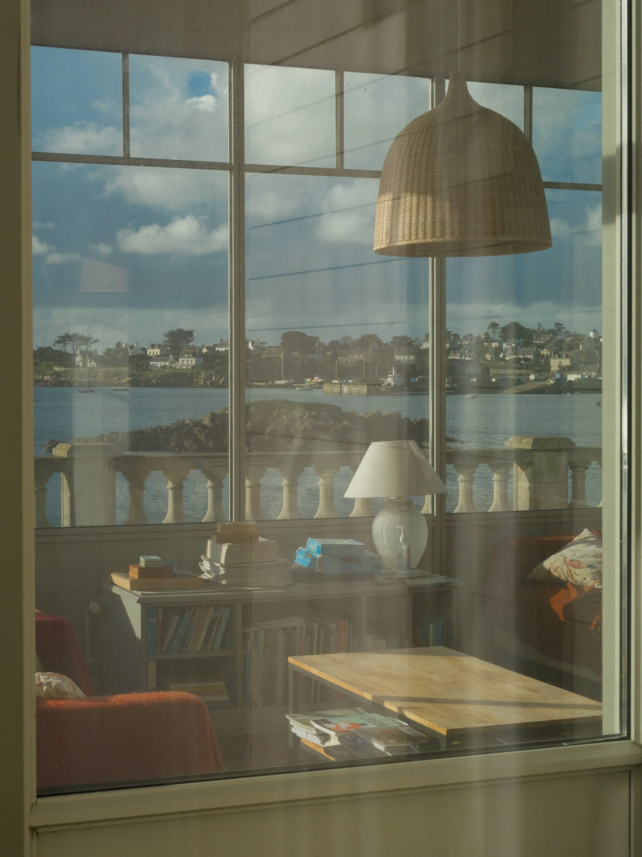 Vue intérieure d'une pièce donnant sur un fleuve avec des maisons, une table, une lampe, un lampadaire en bambou, et un écran de fenêtre avec un cadre blanc.