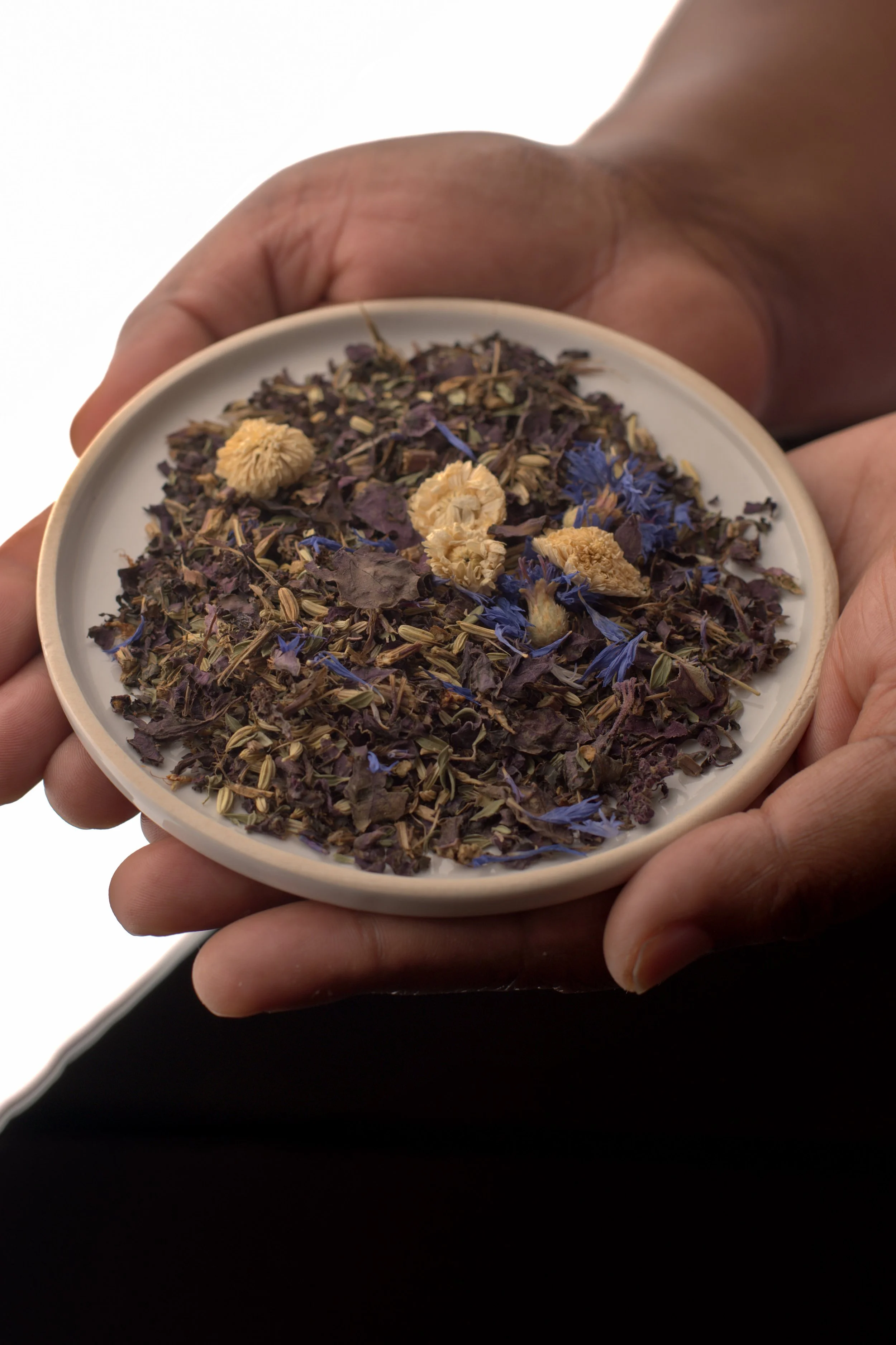 Une main tient un petit plat blanc contenant un mélange de fleurs séchées et de feuilles, avec des fleurs bleues et beige en évidence.