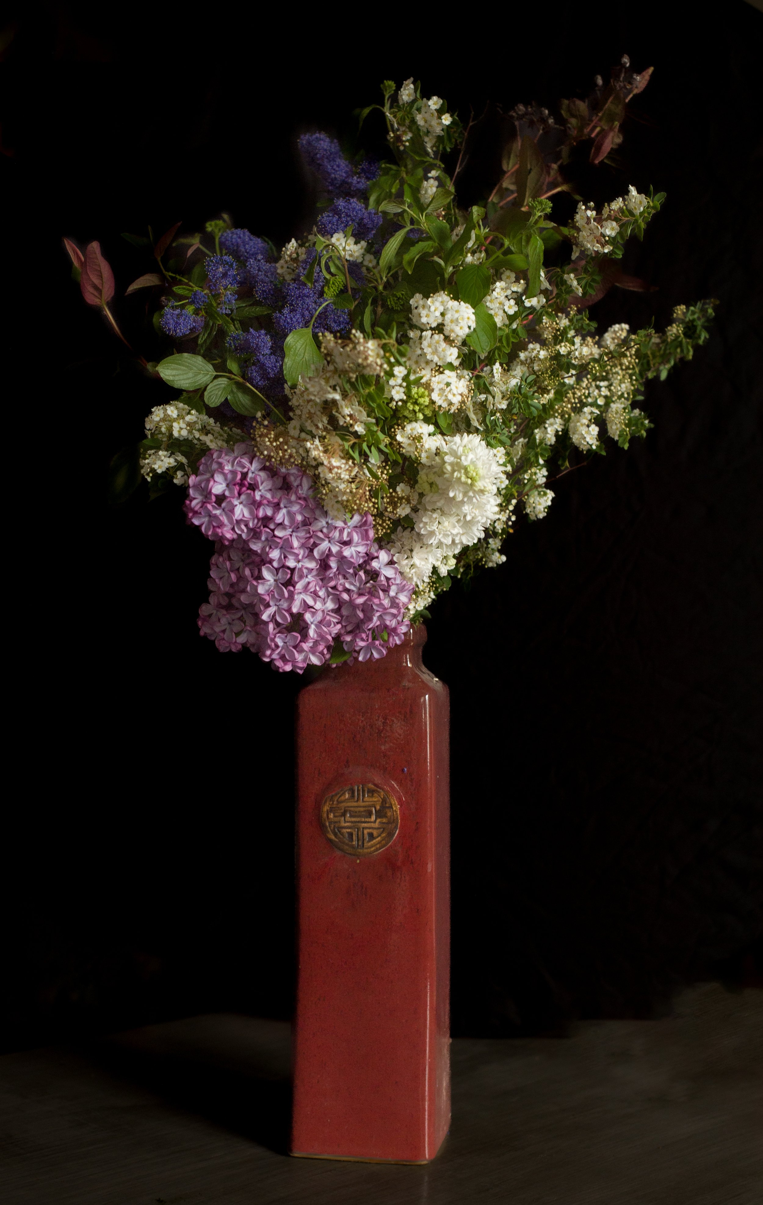 Un vase rouge en céramique avec un motif doré, contenant un bouquet de fleurs colorées, dont des hortensias roses et blanches, et autres fleurs violettes et blanches, contre un fond noir.