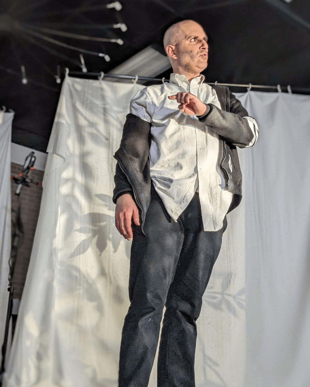 A man with a shaved head standing in front of a white curtain, dressed in a white shirt and dark pants, with a black jacket draped over his shoulders. He is looking to the side with an intense expression.