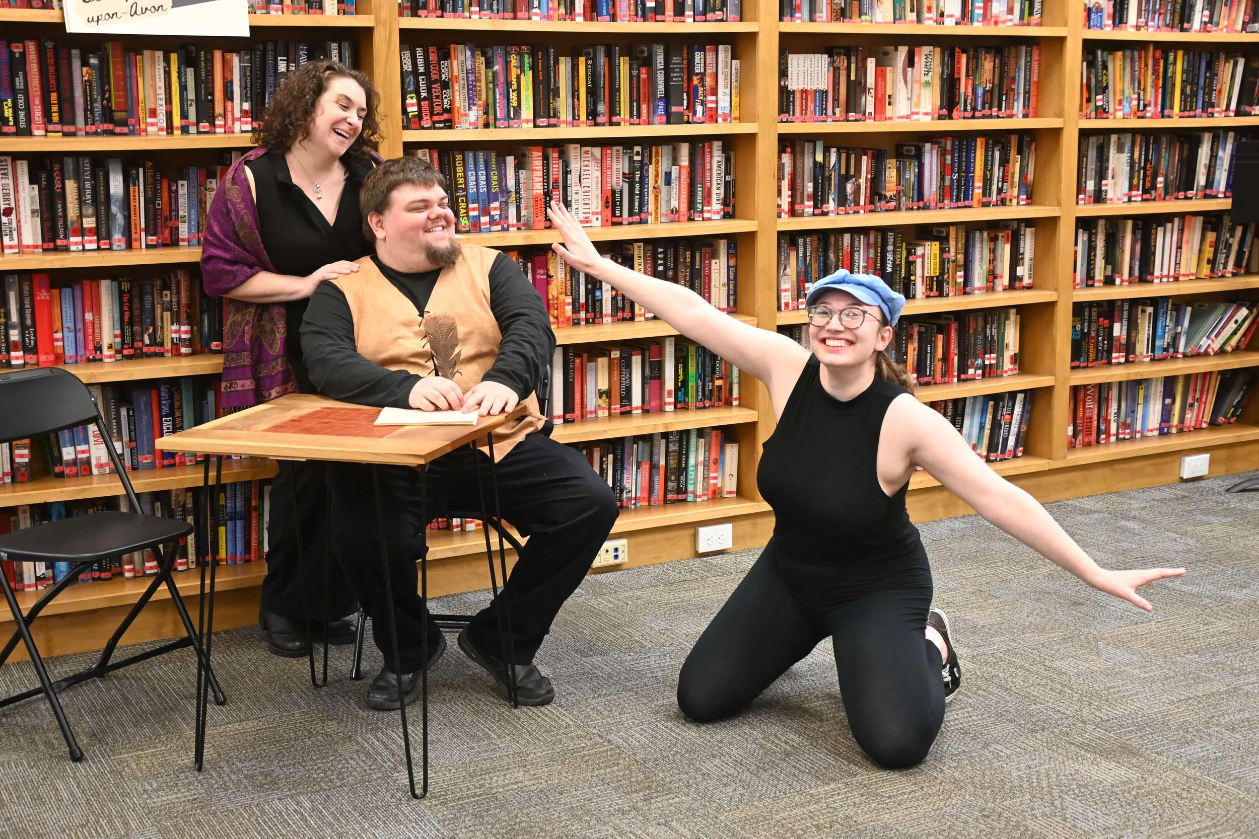 Three people in a library. Two are sitting at a table and one is kneeling in front. They are smiling and laughing, with one woman reaching out towards the man, and a woman standing behind them.