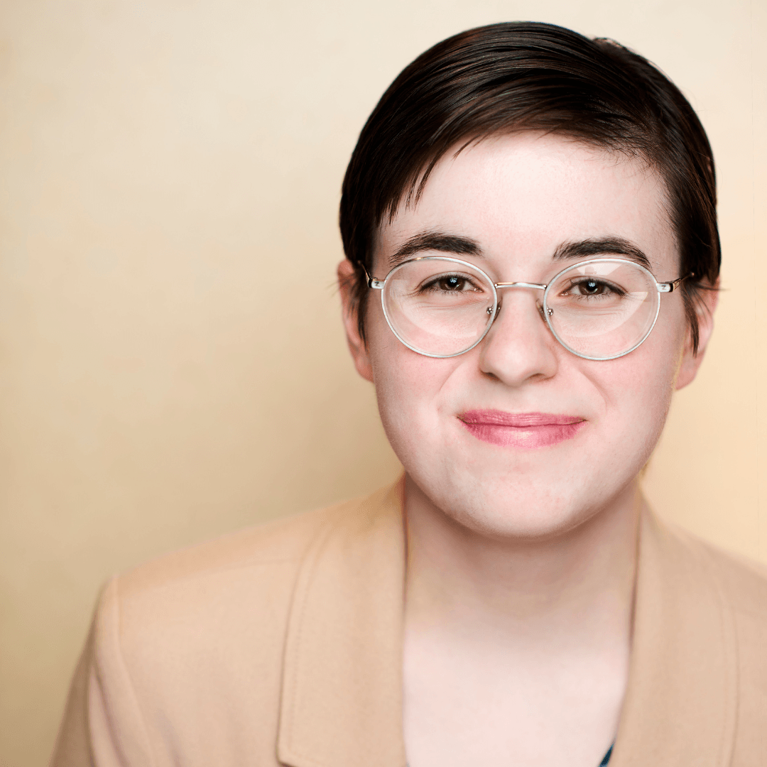 Headshot of an actor with short hair smiling with a closed mouth and wearing round glasses and a tan top 