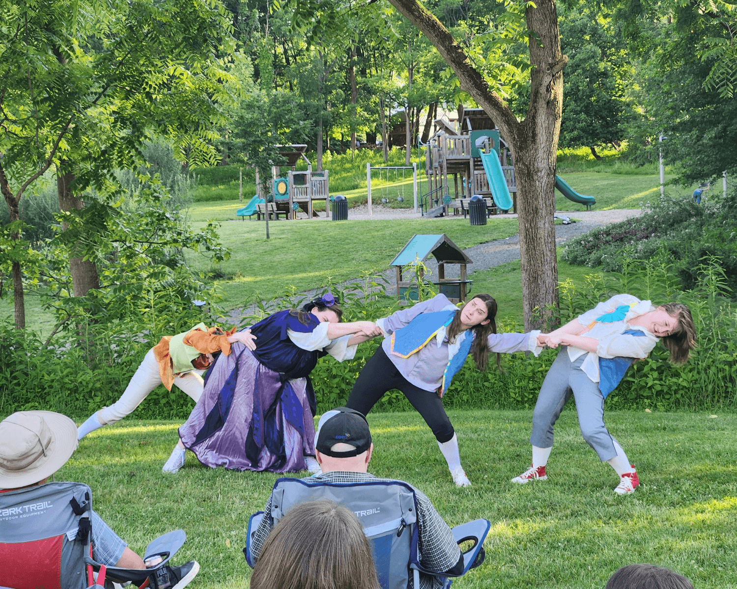 Four women performing a tug-of-war during an outdoor event on a grassy field with trees and a children's playground in the background.