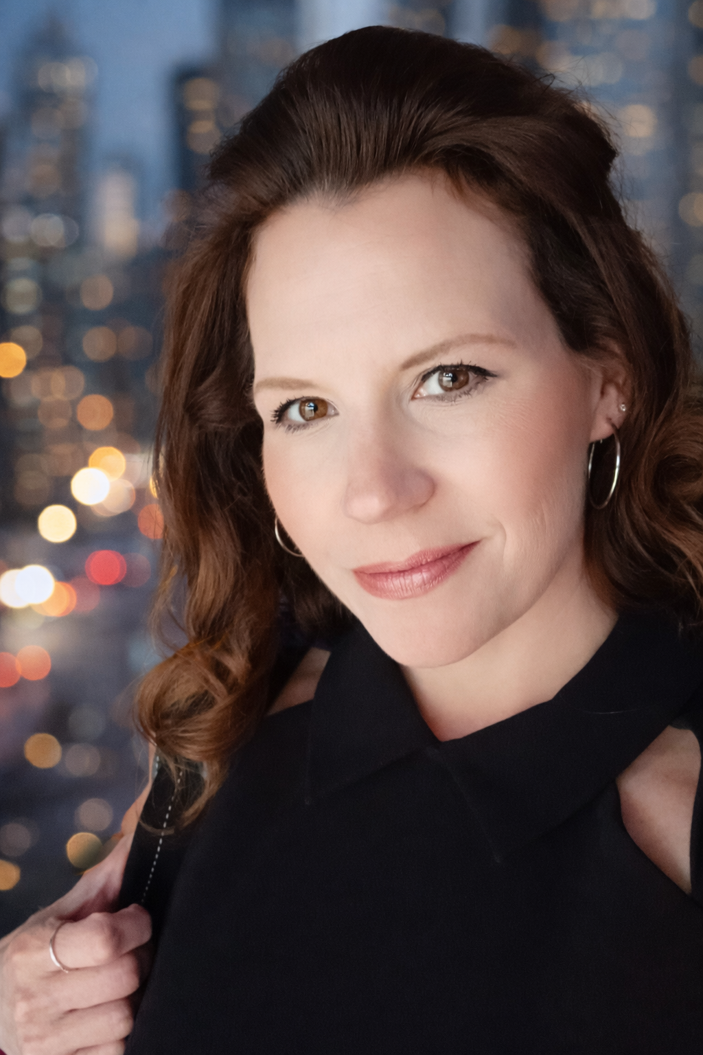 A woman with brown hair and earrings smiling, with city lights blurred in the background.