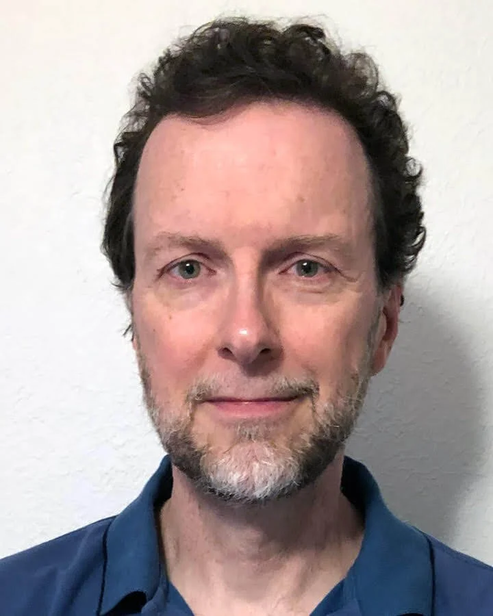 Headshot of a man with short, dark curly hair wearing a dark blue collared shirt against a white wall