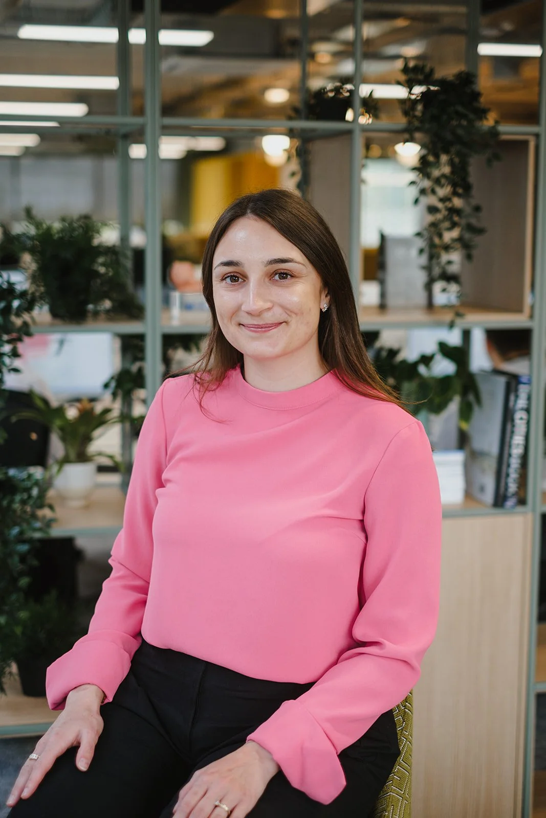 A woman with long brown hair sitting in an office. She is wearing a pink long-sleeved blouse and black pants, and is smiling at the camera. The background features office furniture, plants, and shelving with books and files.