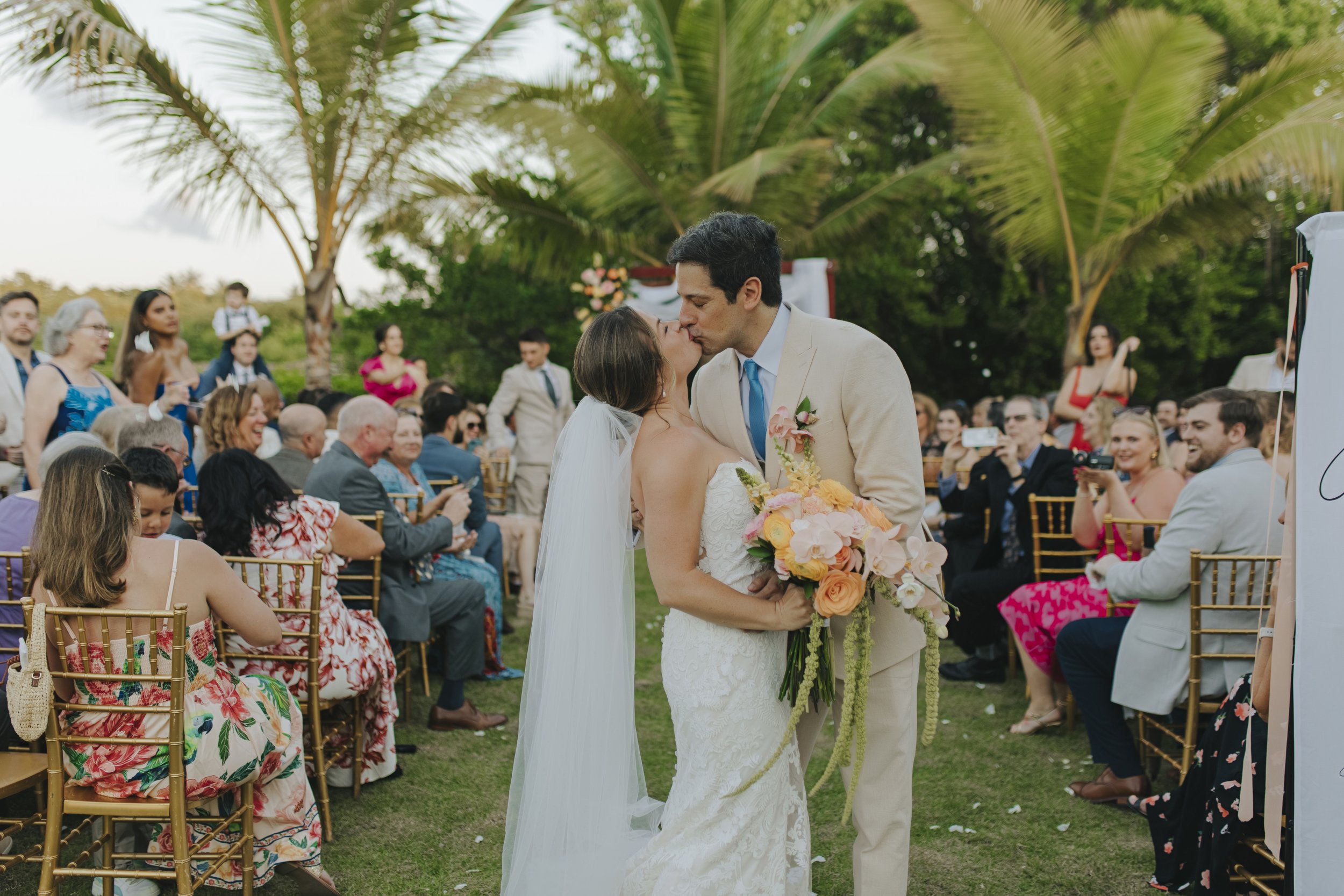 Bride and groom kissing at the end of their garden wedding ceremony in Punta Cana, surrounded by cheering guests under palm trees