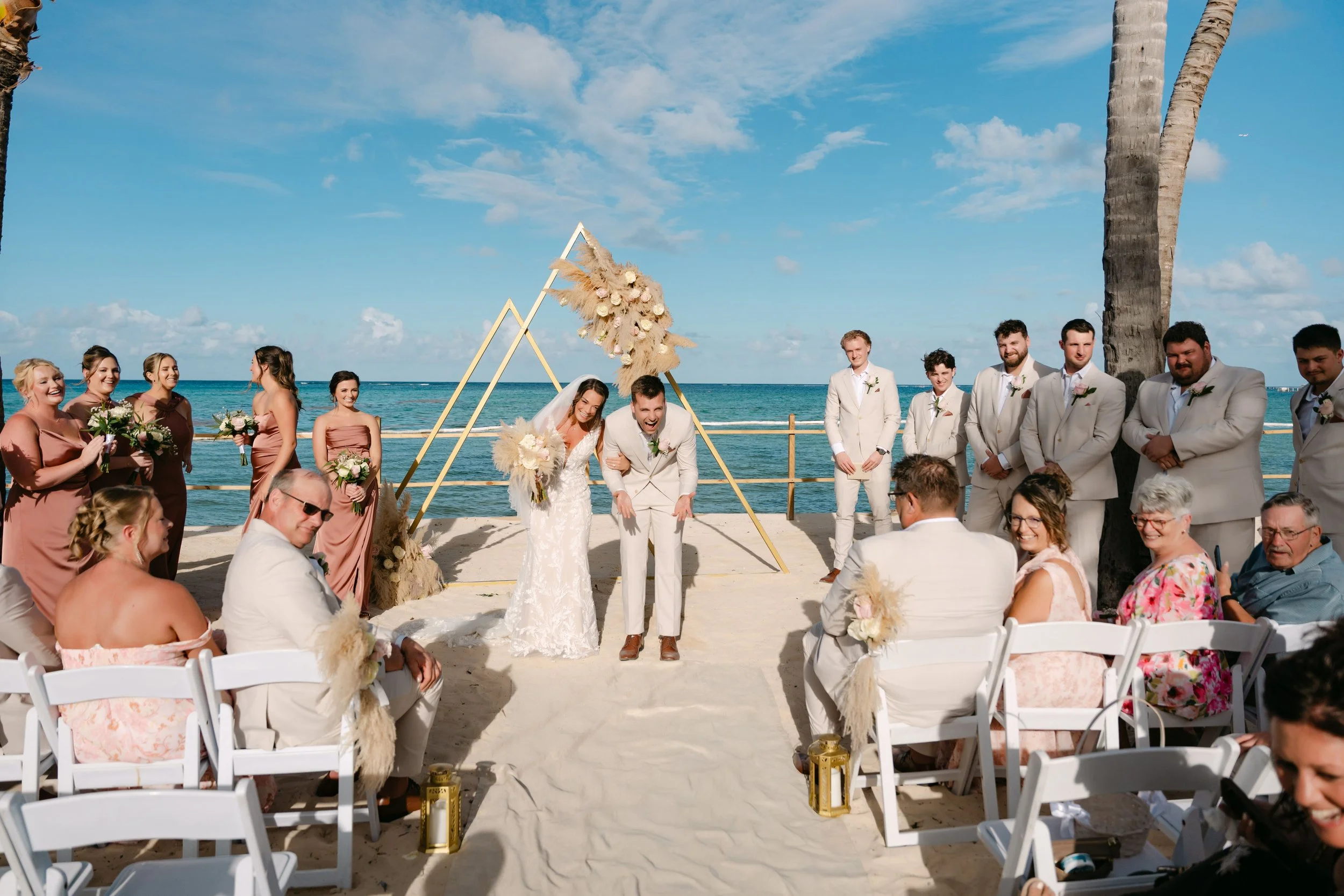 Bride and groom smiling as they walk down the aisle after their beachfront wedding ceremony in Punta Cana, surrounded by family, friends, and a geometric floral arch overlooking the Caribbean Sea.