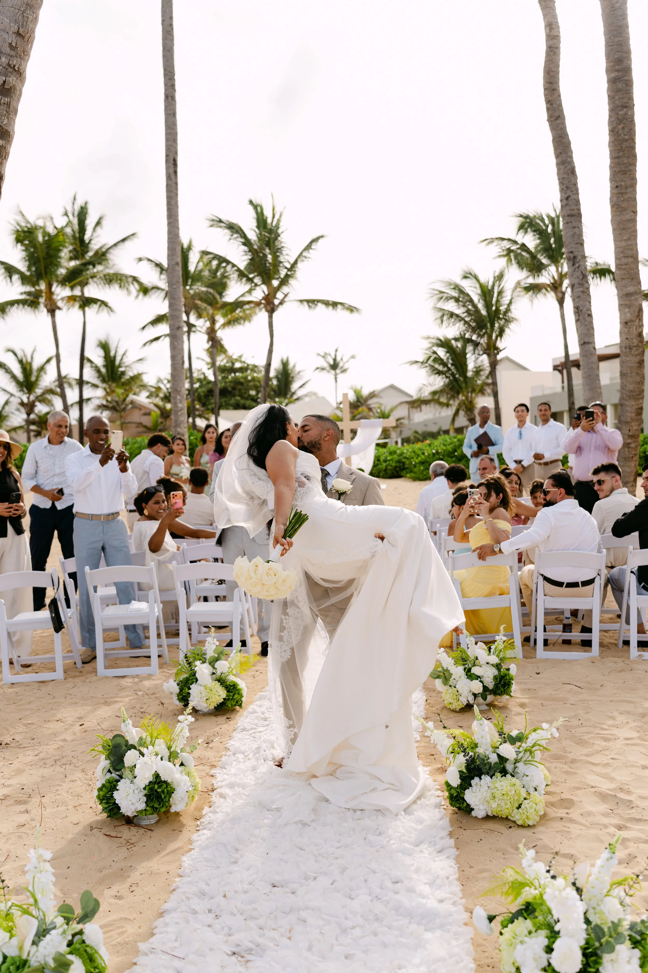 Wide view of the beach ceremony with the couple at the end of a floral aisle, white chairs, guests and palm trees
