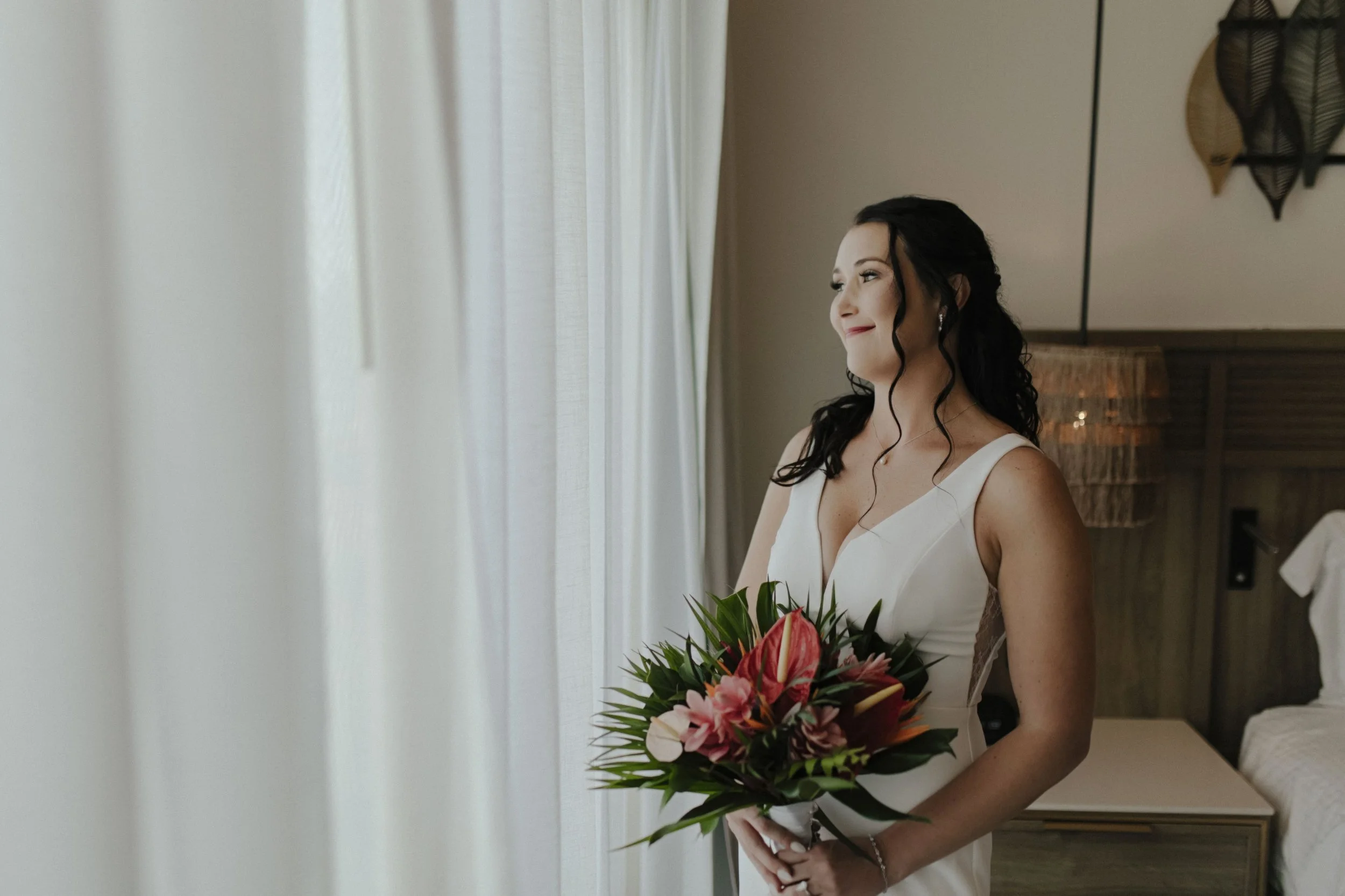 Bride in her wedding gown holding a tropical bouquet by a window in soft natural light at Secrets Tides