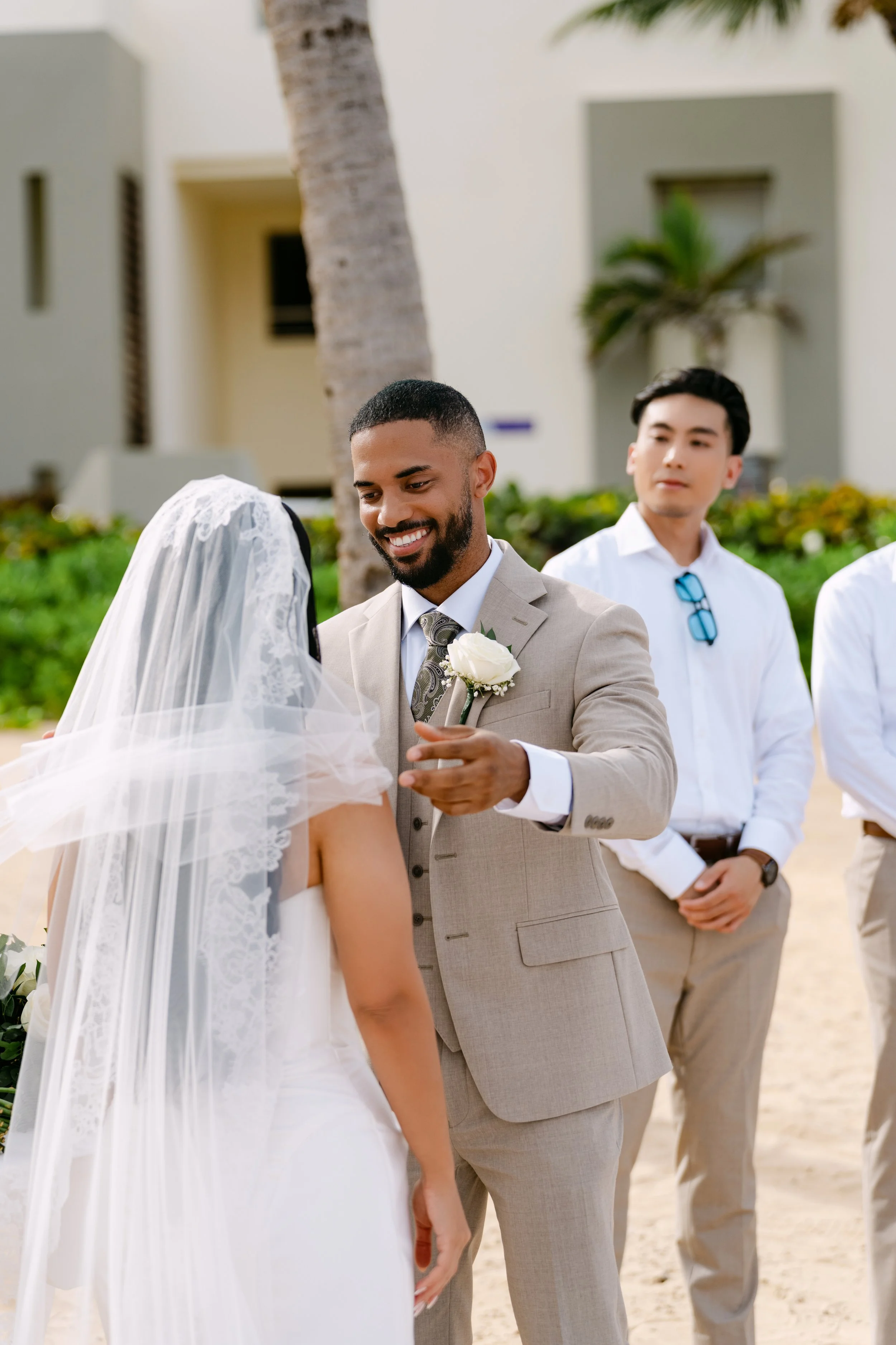 Groom in a tan suit facing the bride in her veil during the beach ceremony with palm trees behind them