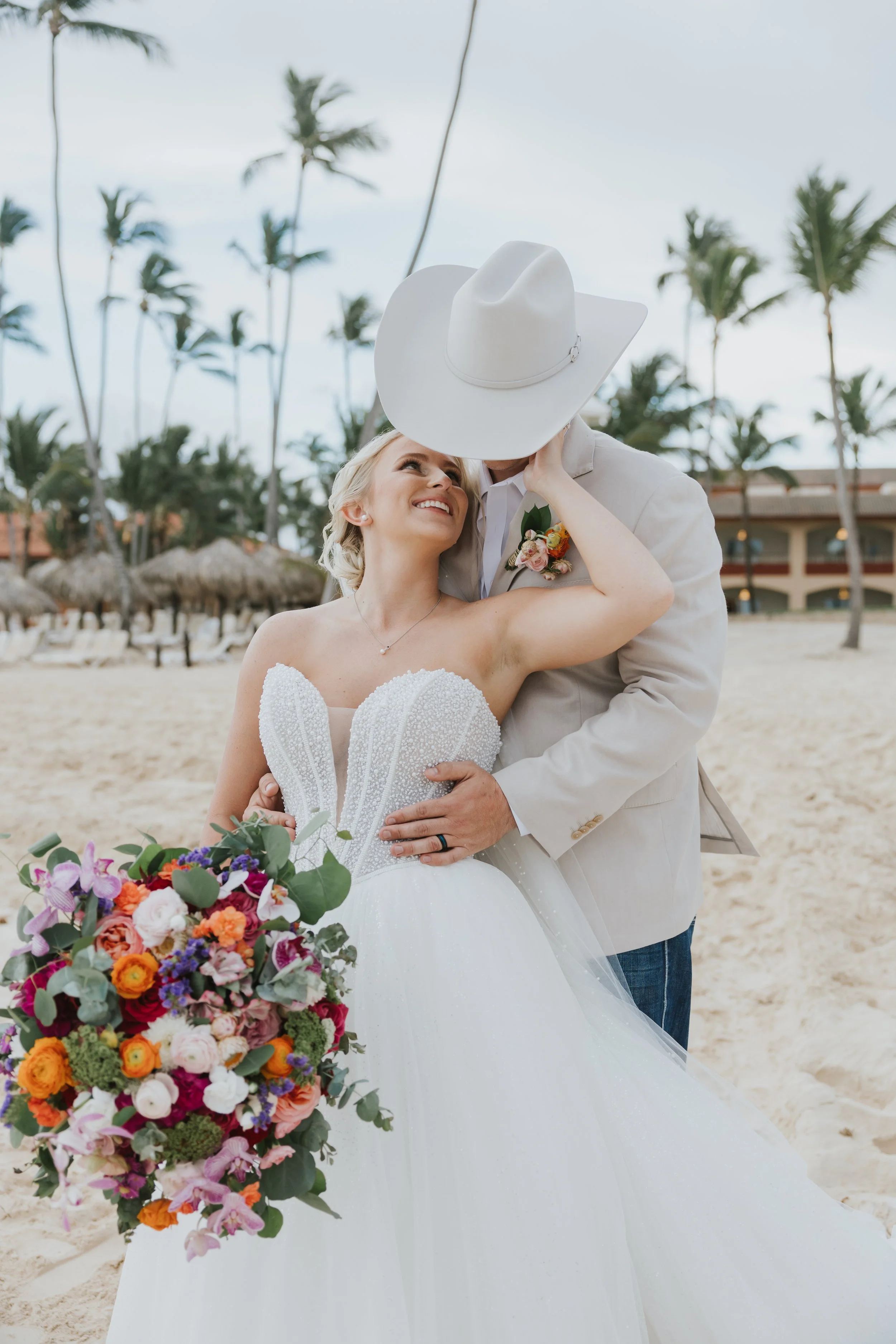 Close-up portrait of bride in a beaded sweetheart bodice holding her jewel-tone bouquet, groom in cowboy hat embracing her, palm trees and beach umbrellas in the background at Majestic Elegance Punta Cana
