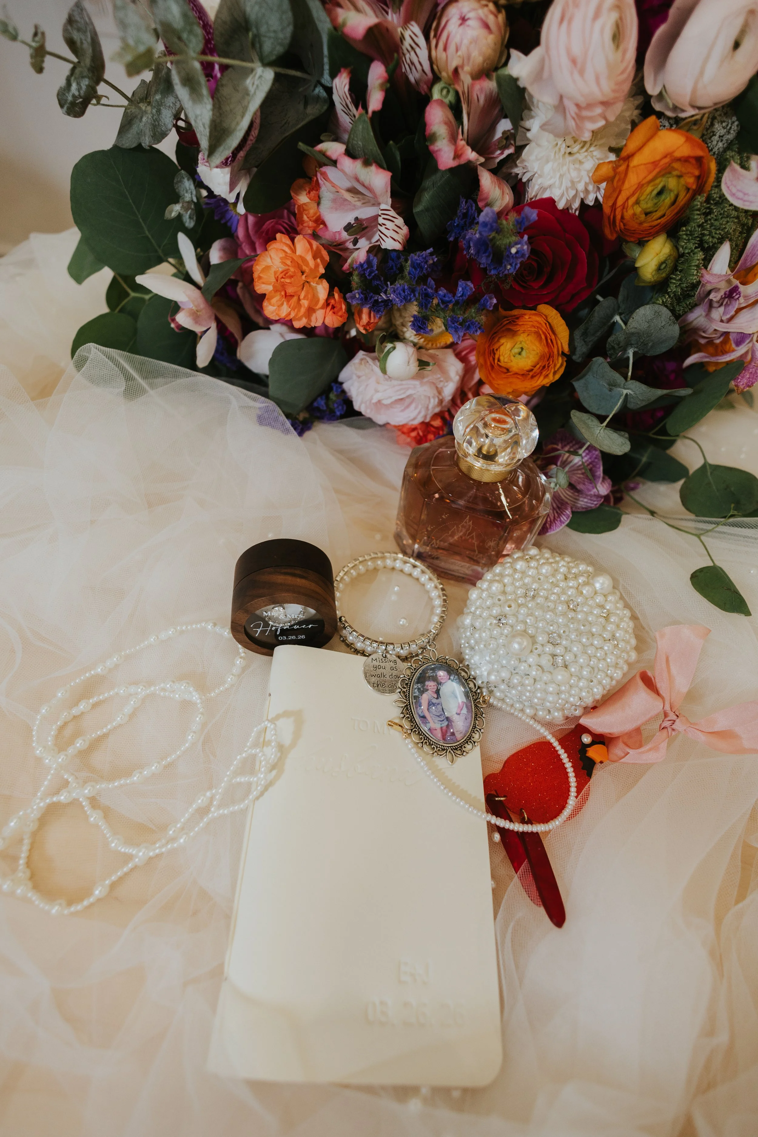 Wedding day details flat lay with bridal veil, jewel-tone bouquet, perfume bottle, pearl bracelets, engraved ring box, and a locket photo tied with ribbon at Majestic Elegance Punta Cana