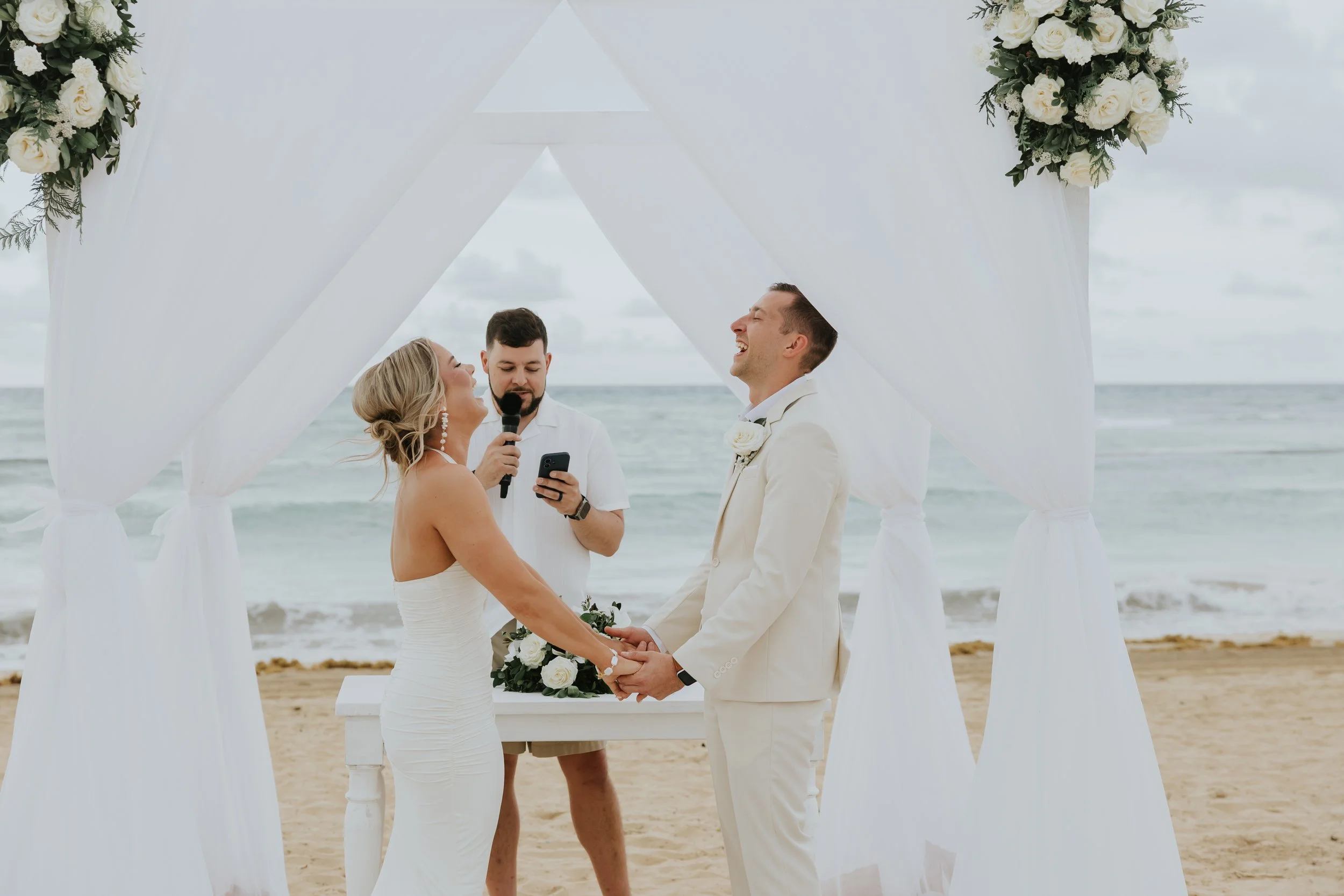 Bride and groom holding hands and laughing under a white draped arch with white floral arrangements, with the officiant and the ocean behind them at a Secrets Tides beach wedding