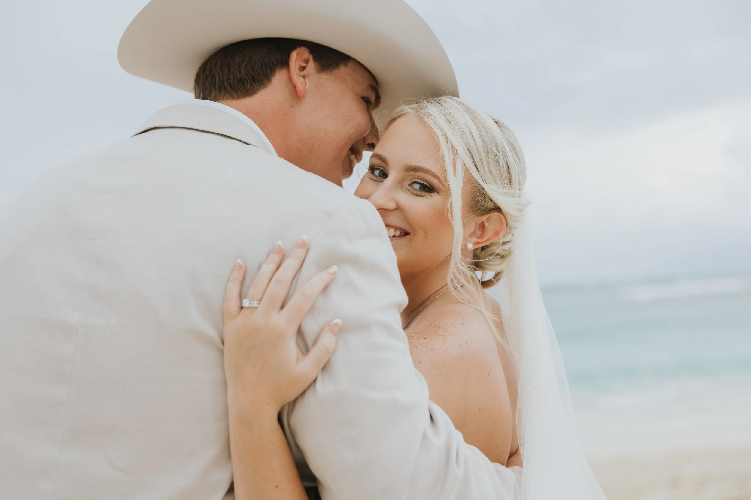 Bride and groom laughing forehead to forehead in a candid beach portrait, groom wearing a cream blazer and cowboy hat, at Majestic Elegance Punta Cana