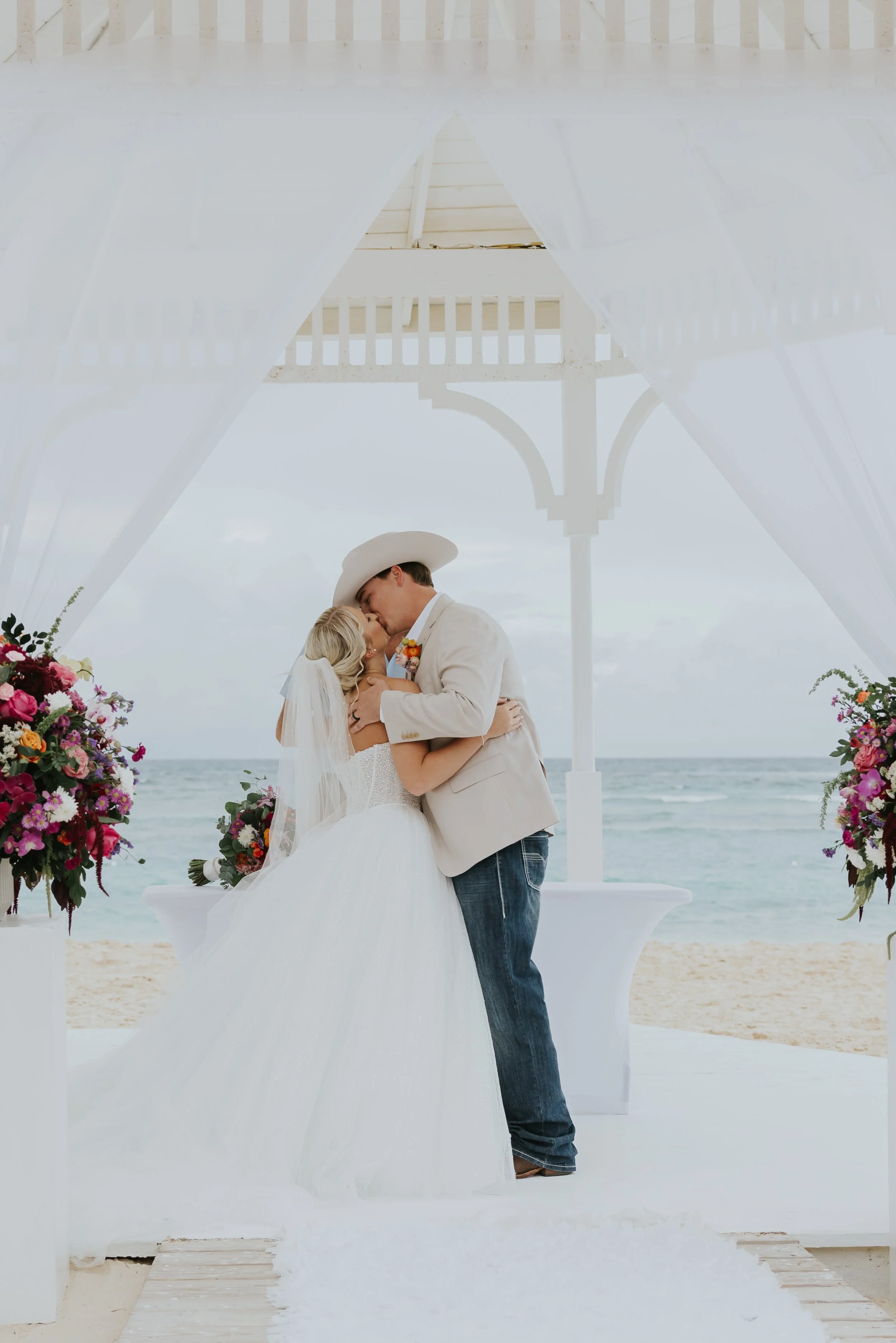 Bride and groom in a cowboy hat share their first kiss at the beachfront gazebo altar, jewel-tone floral arrangements on either side, at Majestic Elegance Punta Cana