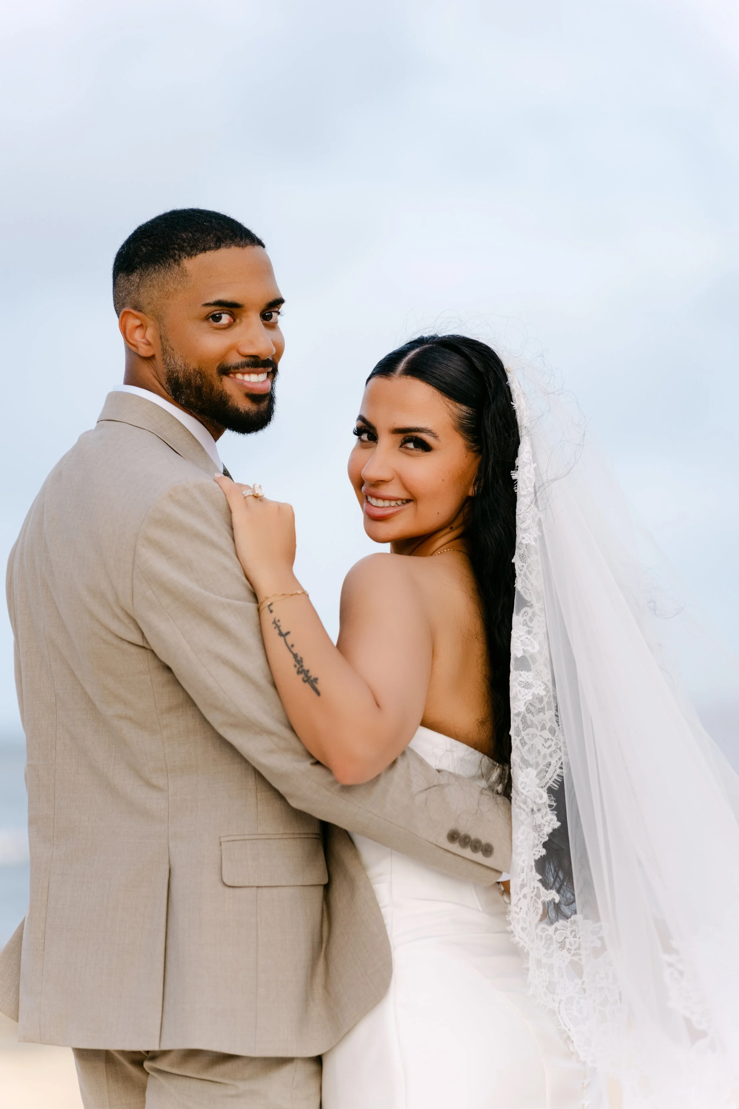 Bride in a lace-edged veil and groom in a tan suit embracing and smiling at the camera against a soft sky