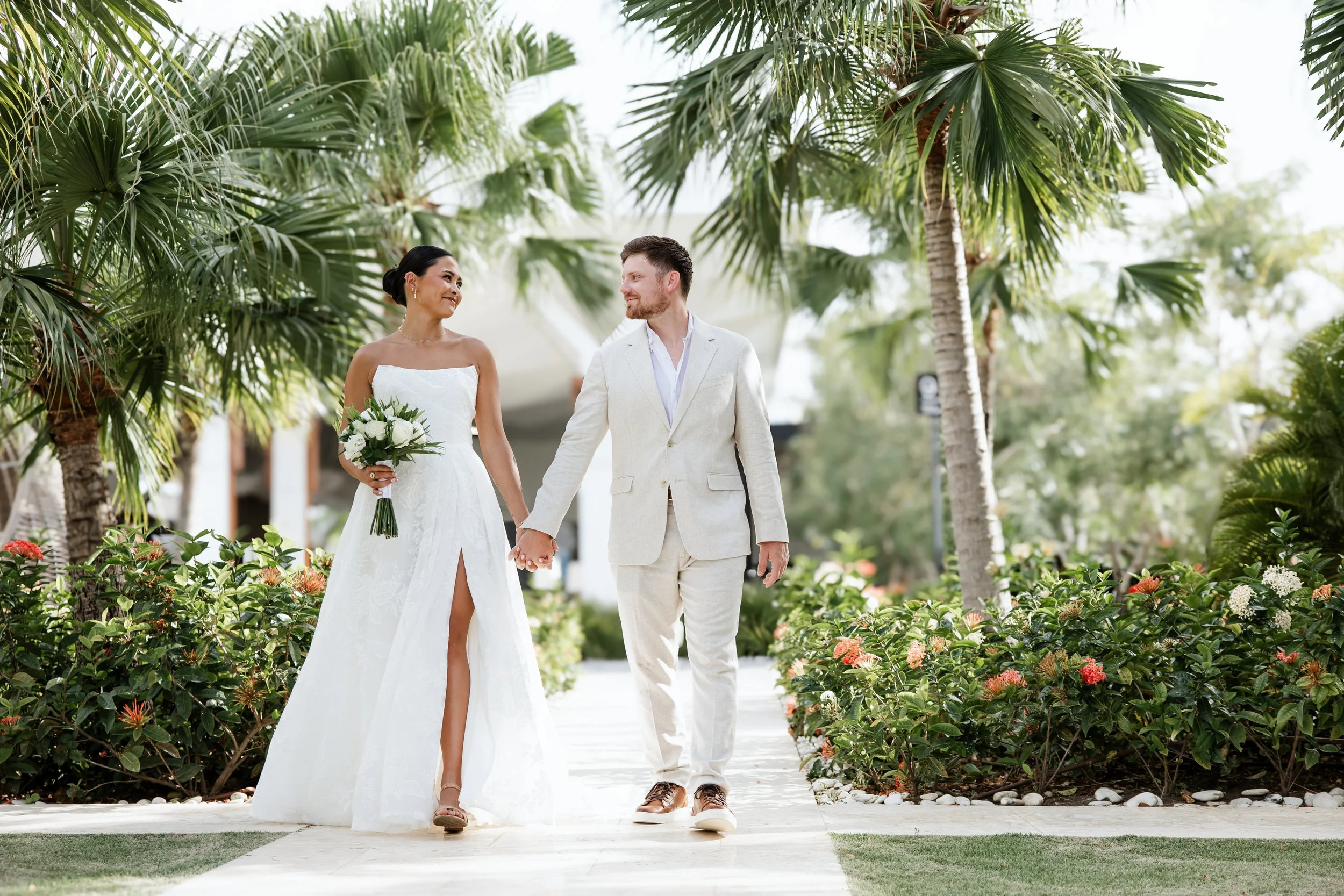 Bride in a white lace gown with a high slit holding a white rose bouquet and groom in a cream linen suit walking hand in hand and smiling at each other along a garden path lined with palm trees and orange ixora flowers at Secrets Tides Punta Cana